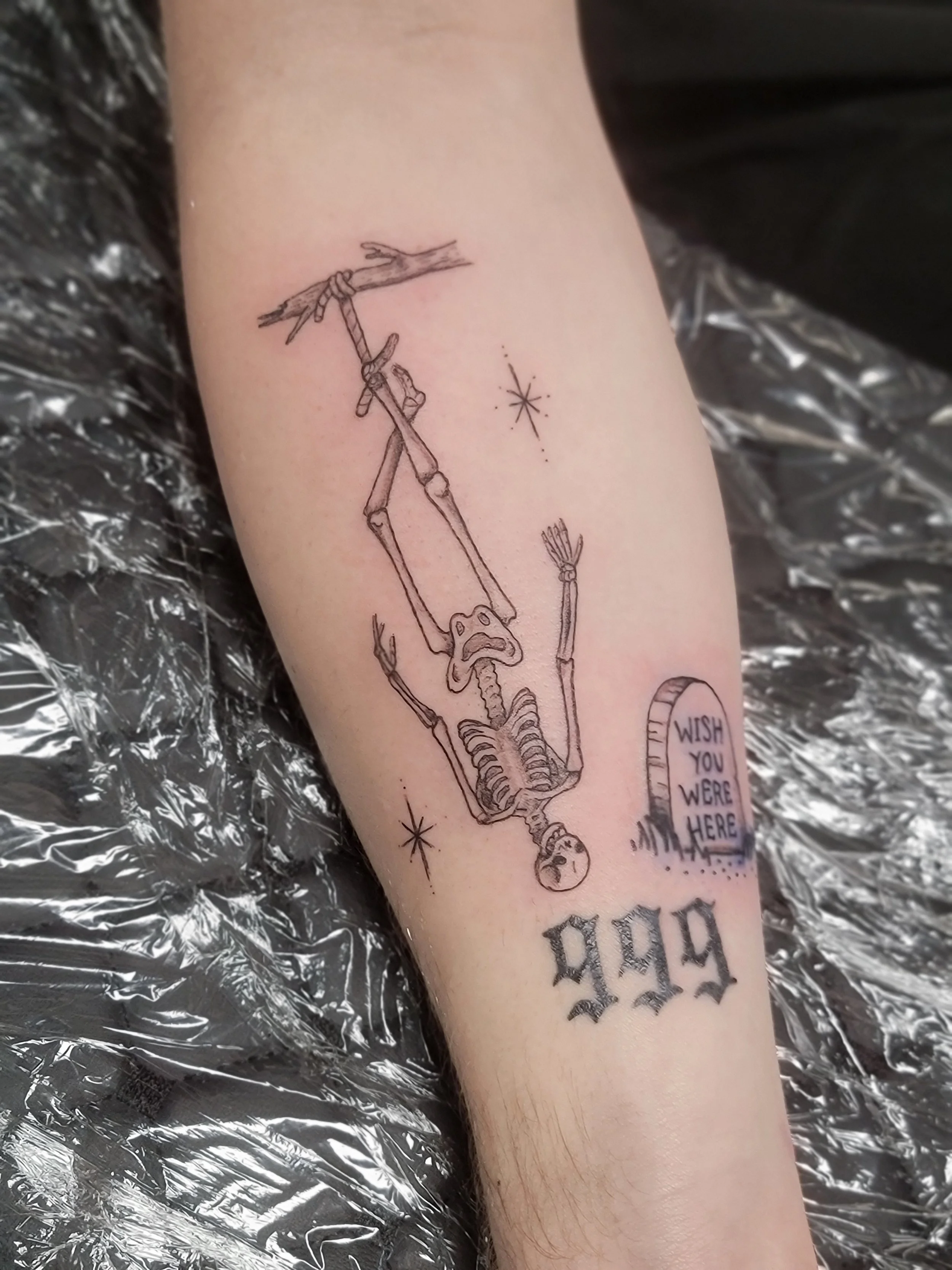 A tattoo of a skeleton hanging from a tree branch, with a small skull at the bottom and stars around it. Part of a speech bubble says 'WISH YOU WERE HERE'.