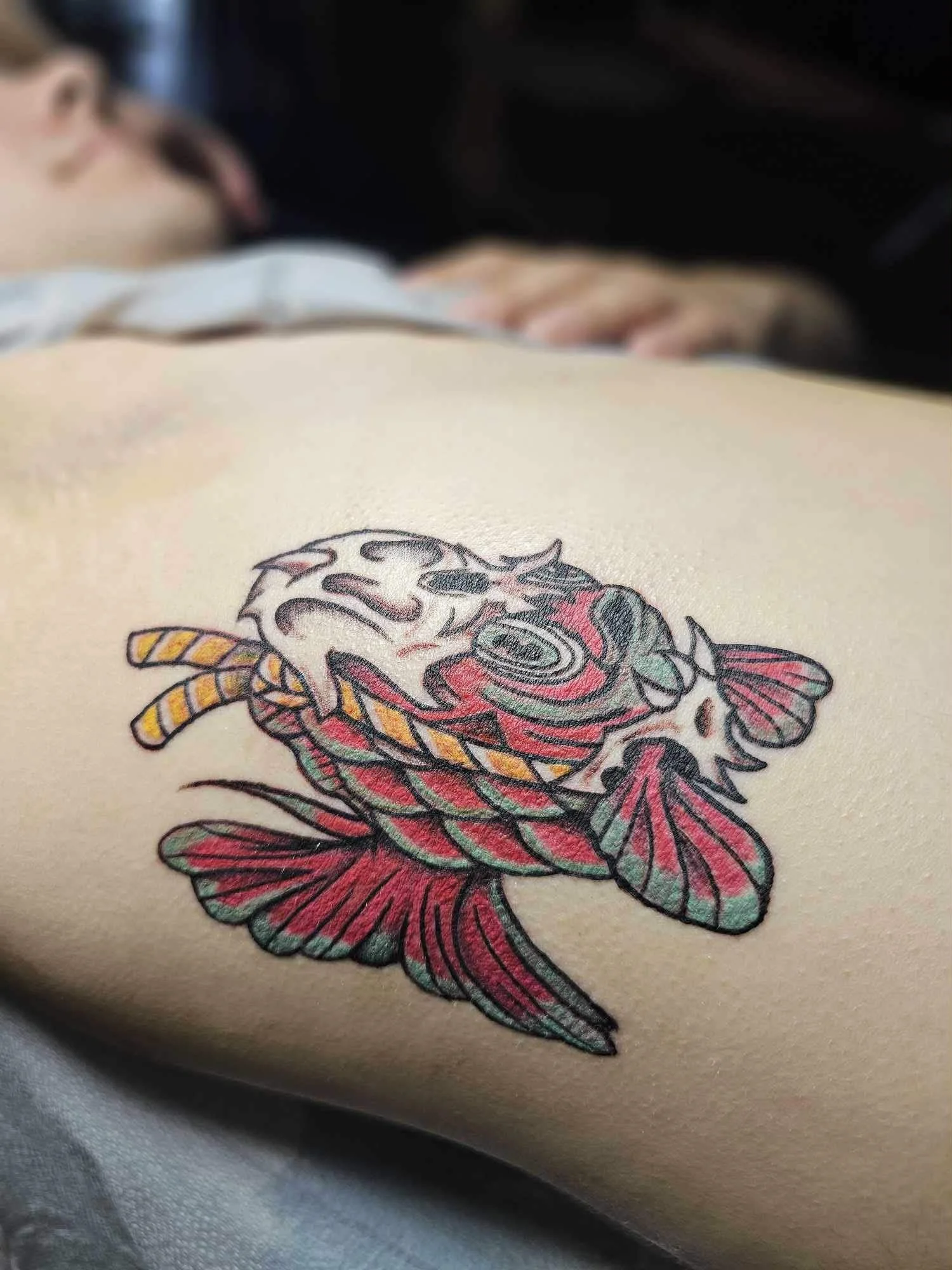 Tattoo of a fish with a skull on the body, colored in red, black, yellow, and green, on a person's upper arm.
