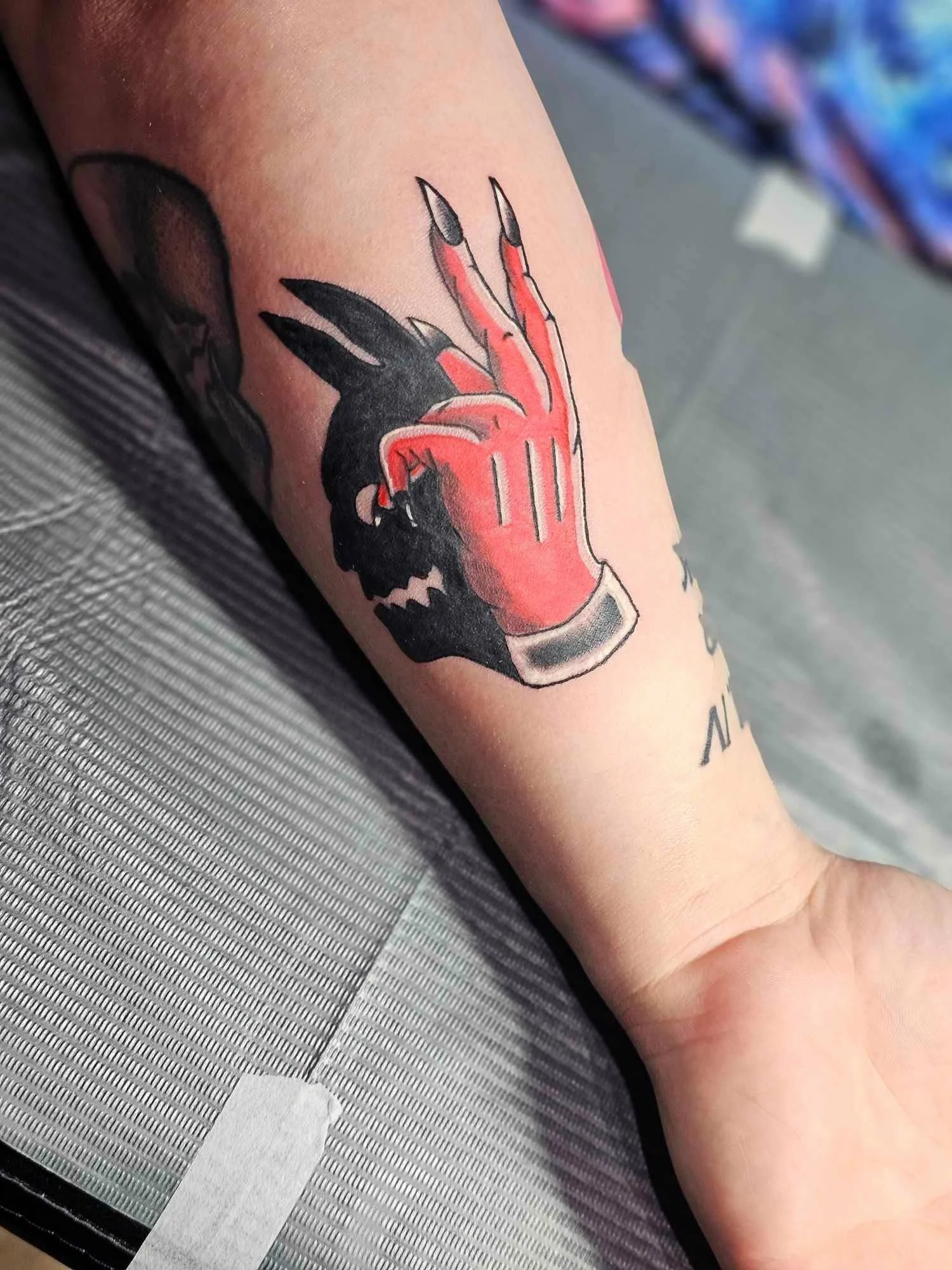 Tattoo of a red and black cartoon monster with sharp teeth and claws, making a hand gesture, on a person's forearm.