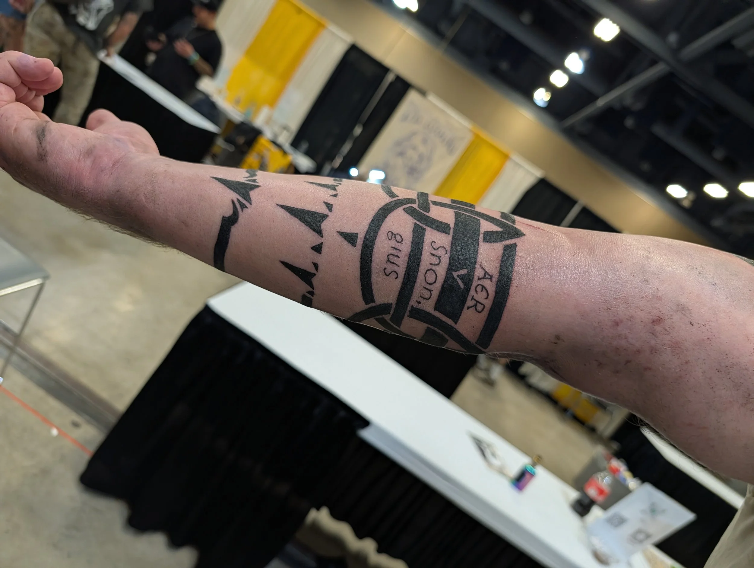 Close-up of a person's arm with a tattoo of a circular emblem and text, in front of a background of store shelves with various products.