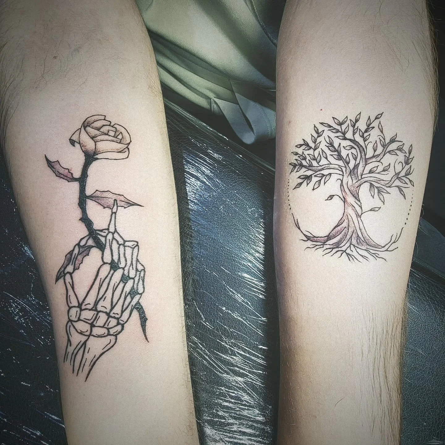 Tattoo of a skeletal hand holding a rose on the left arm and a tree with roots and leaves outline on the right arm.