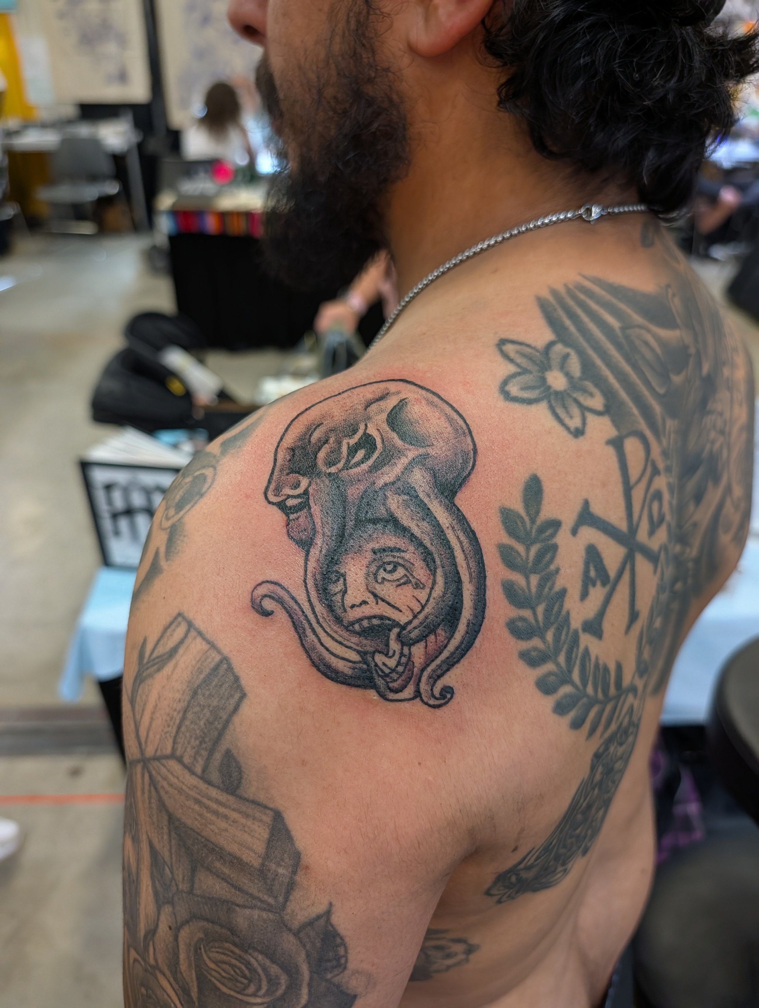 Close-up of a man's shoulder and upper arm tattoo featuring a skull with a snake emerging from its mouth, a woman with a snake in her mouth, and floral and geometric designs in black and gray ink.