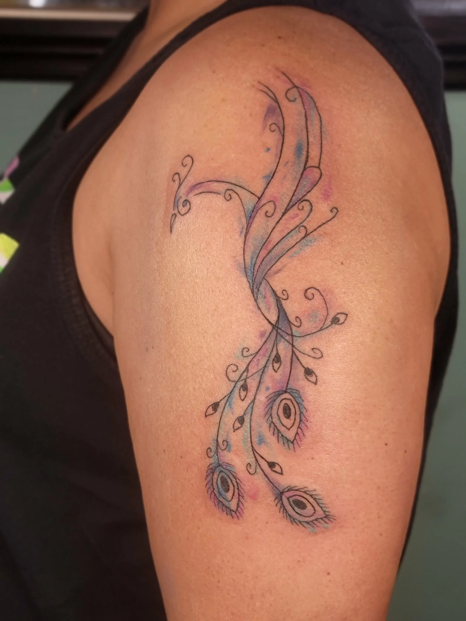 Colorful tattoo of peacock feathers with swirling lines and blue, purple, and black accents on a person's upper arm/shoulder in watercolor style.
