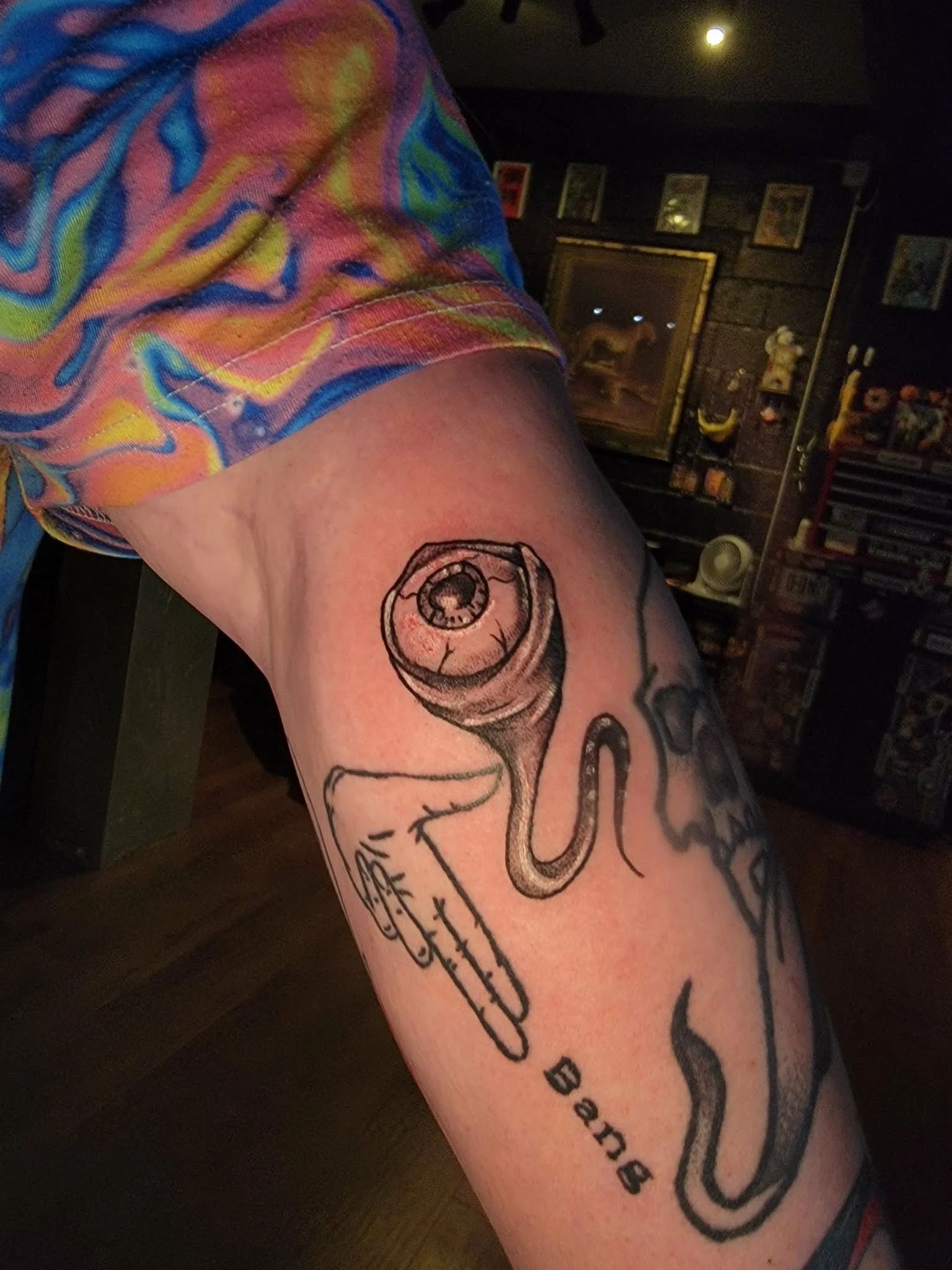 Tattoo of a snake with an eyeball on a person's upper arm, with the word 'Bang' underneath, partially visible, and colorful clothing sleeve.