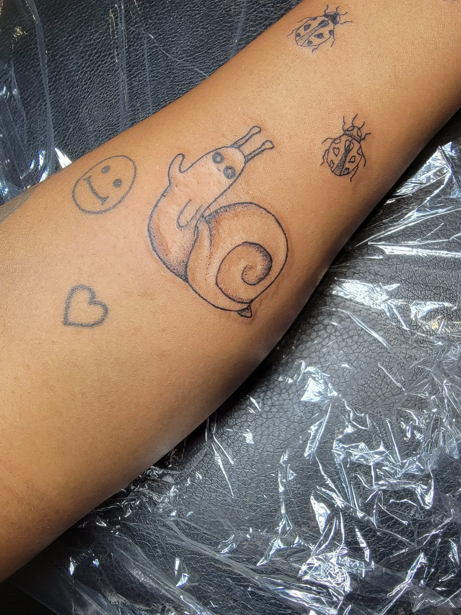 A tattooed arm with a cartoon snail, a smiling face, a heart, and two ladybugs, on a background of plastic and black fabric.