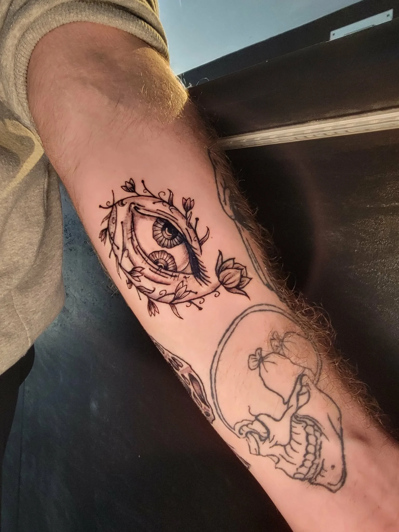 Tattoo of an eye surrounded by leaves on the forearm, and a skull with a bow on the wrist.