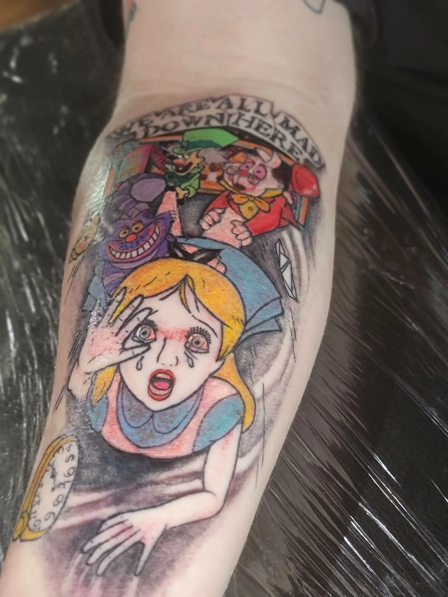 Colorful tattoo featuring characters from Alice in Wonderland, including the Mad Hatter, the Cheshire Cat, and Alice with tears and a distressed expression.