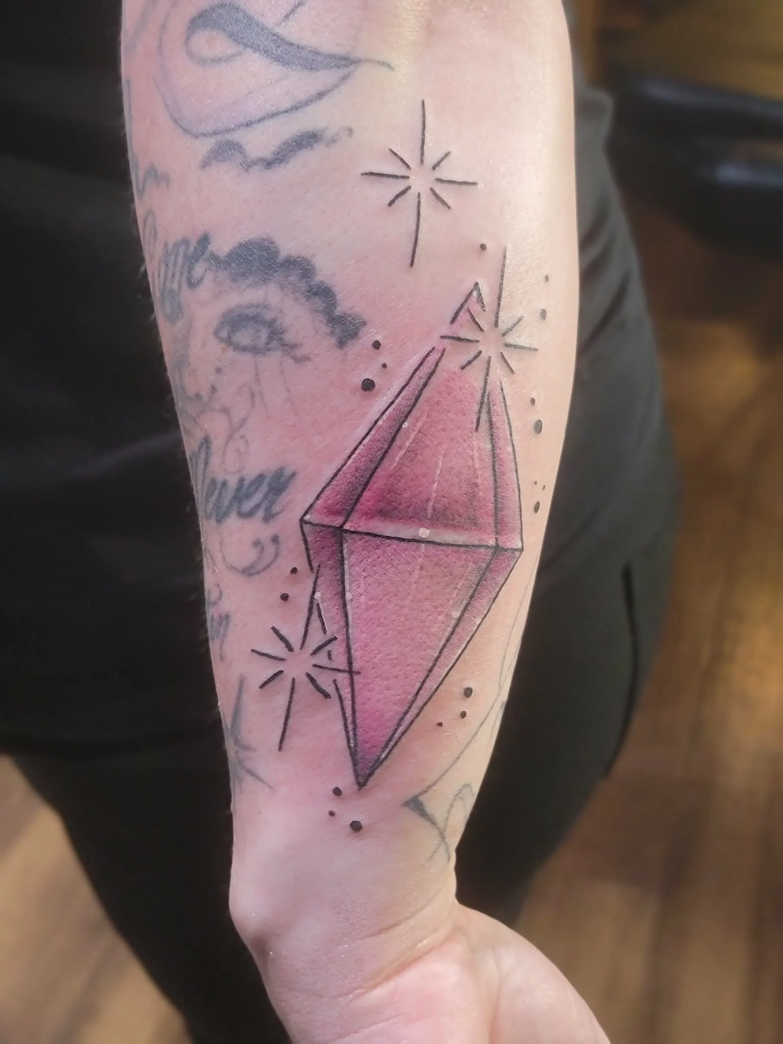 Tattoo of a pink crystal with black outline and shading, surrounded by small stars and dots on a person's forearm.