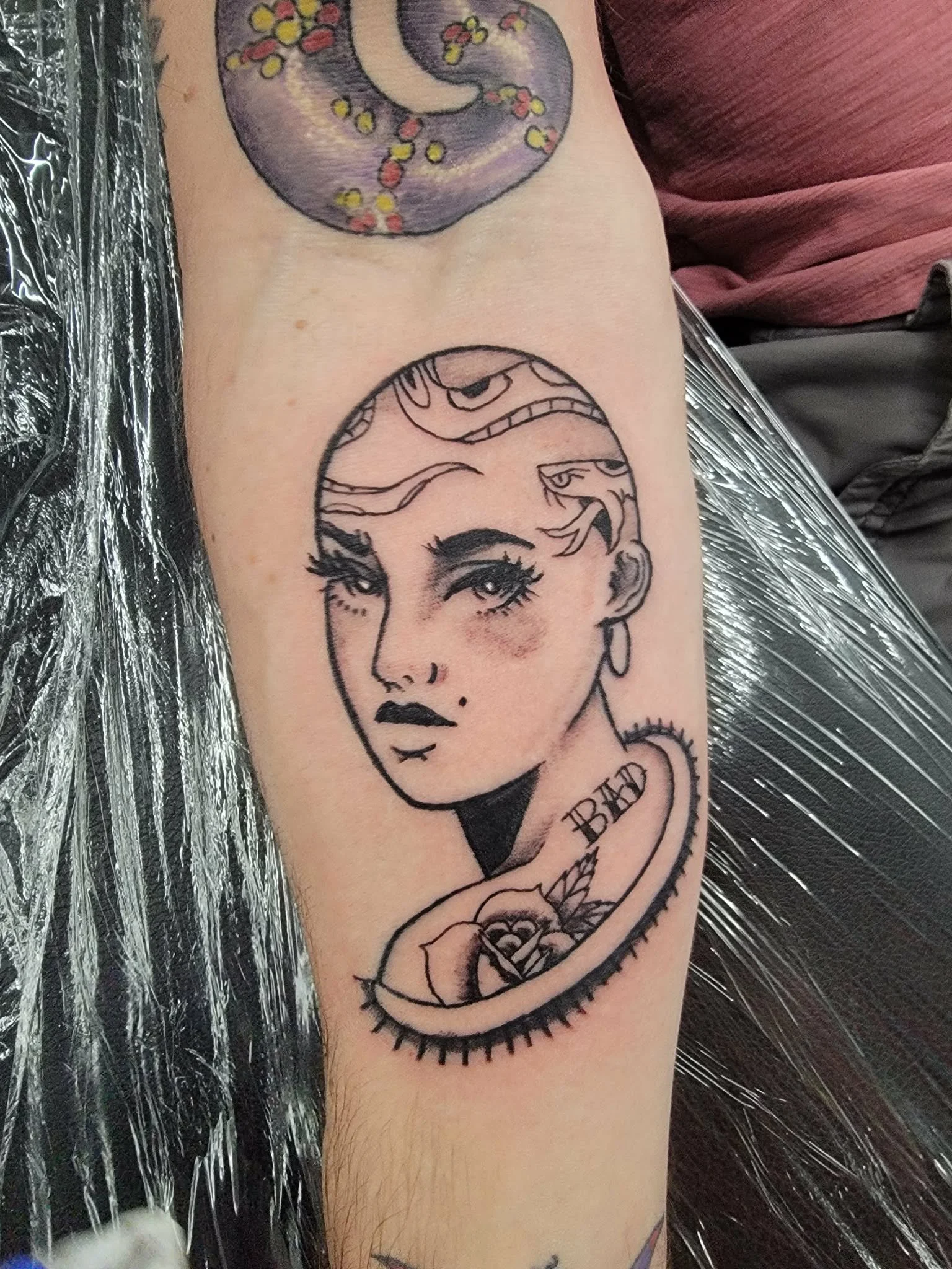 Tattoo of a stylized woman's face with eyeliner, blush, and lipstick, framed by a snake and a rose. The tattoo also has the word "BAD" etched near the snake and some overhead designs. The person is sitting on a surface covered with plastic.
