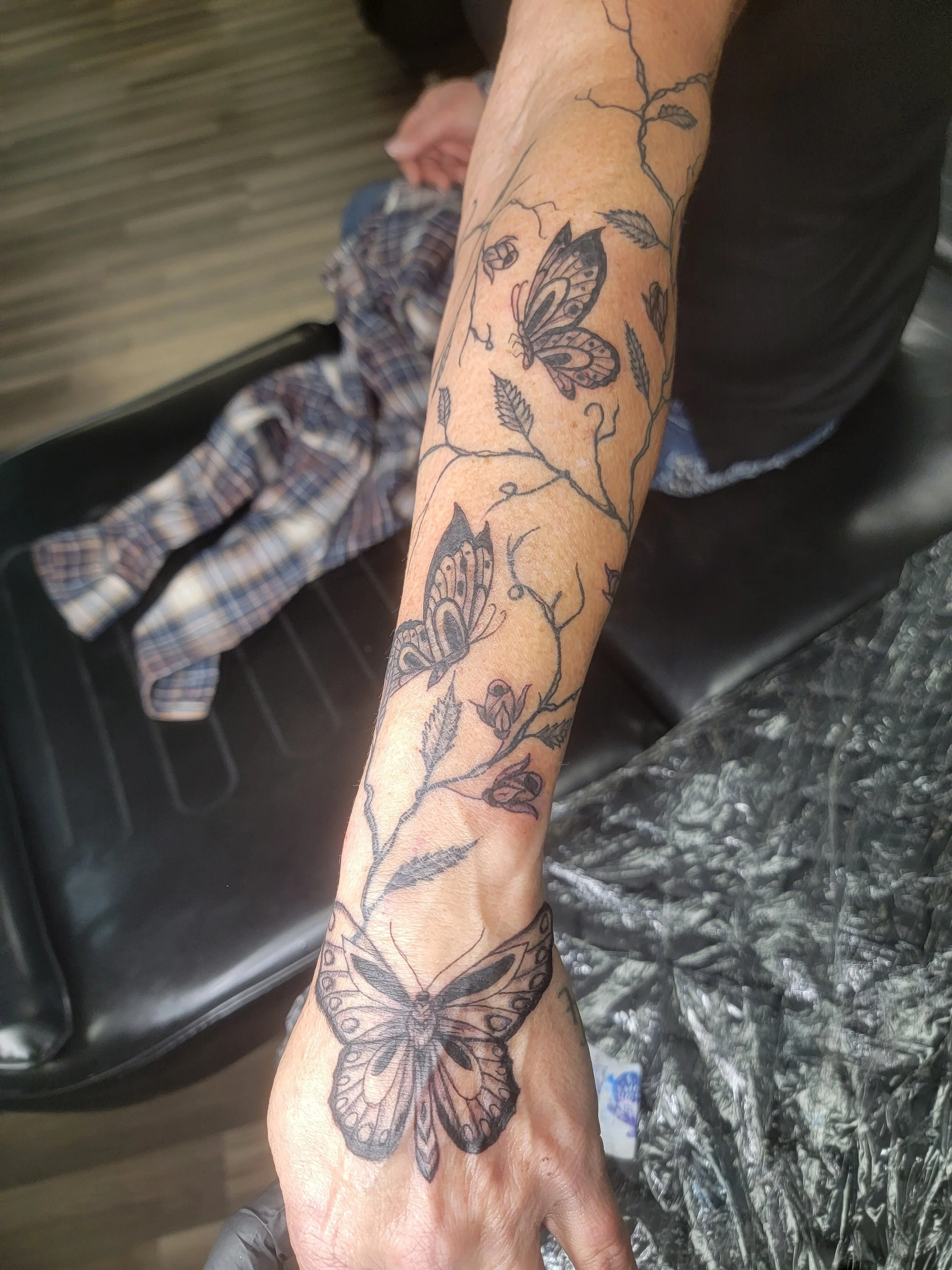 A person's forearm with a black ink tattoo of a butterfly and floral design.