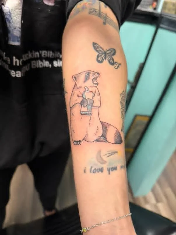 Tattoo of a baby raccoon holding a redbull on a person's forearm, accompanied by a butterfly.