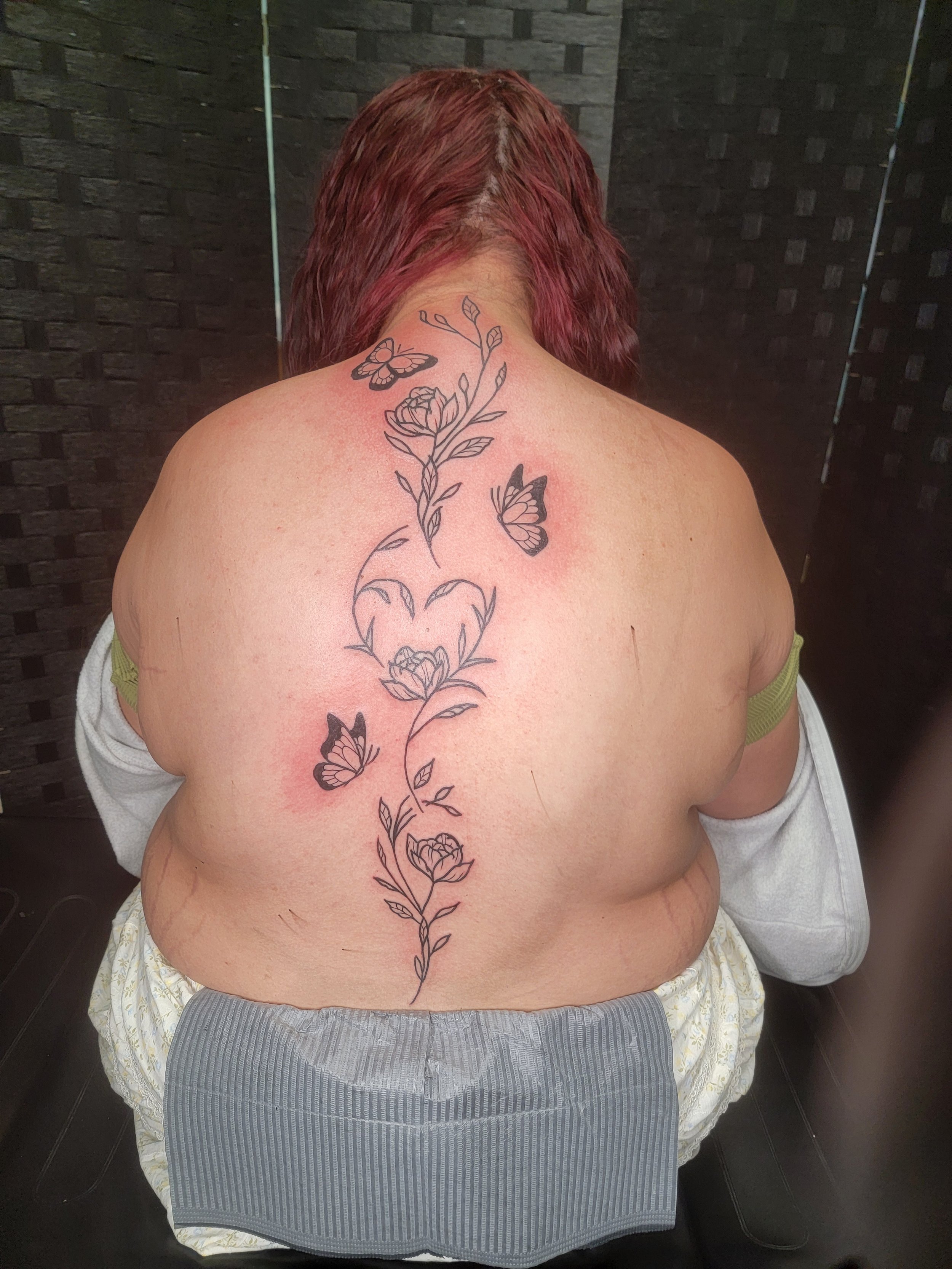 Back of a person with red hair, sitting in a chair, with a tattoo of flowers and butterflies running from the neck to the lower back.