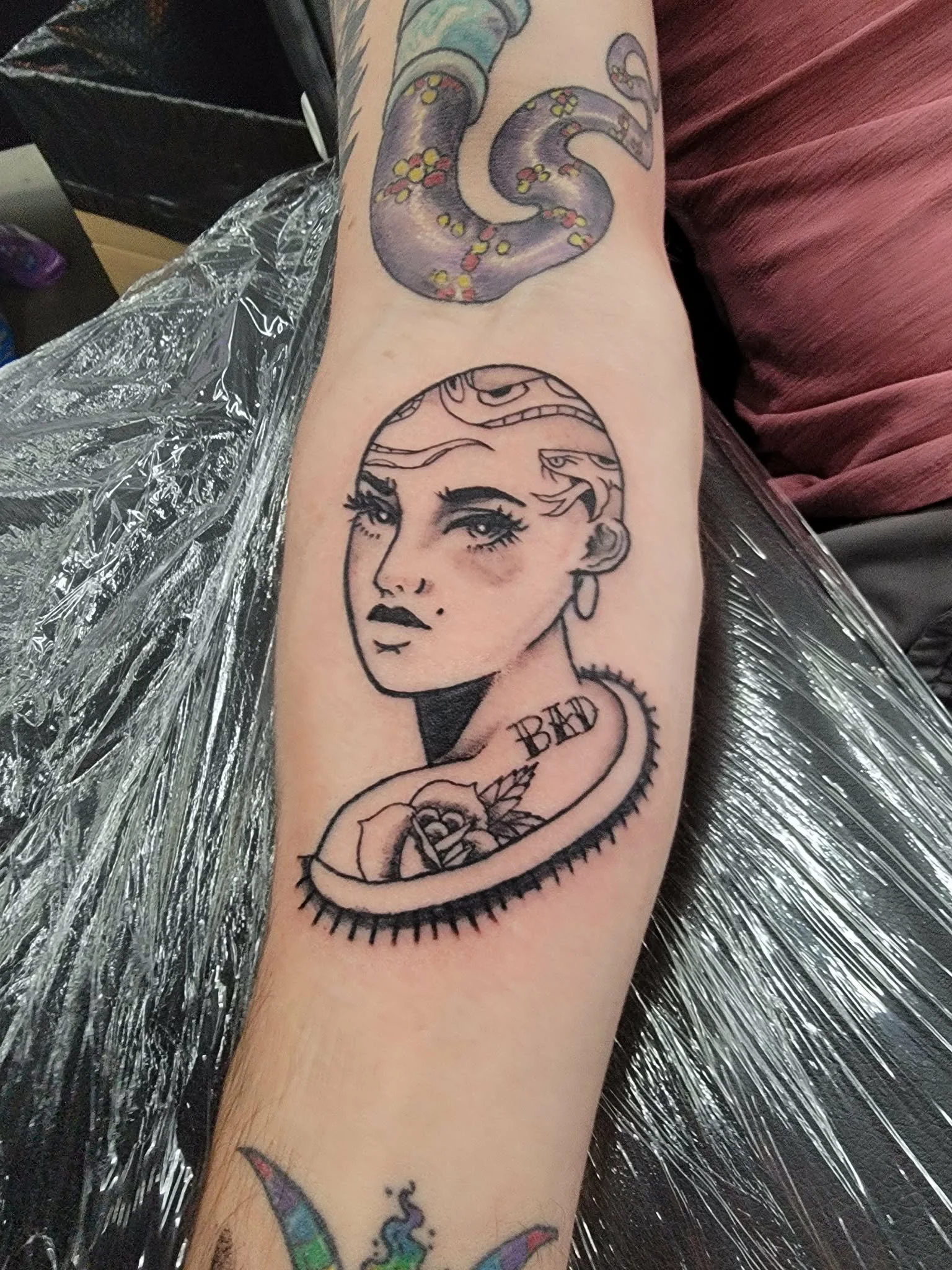 Tattoo of a woman's face with makeup, earrings, and a tattooed headscarf, accompanied by a rose and the word "BAD" underneath.