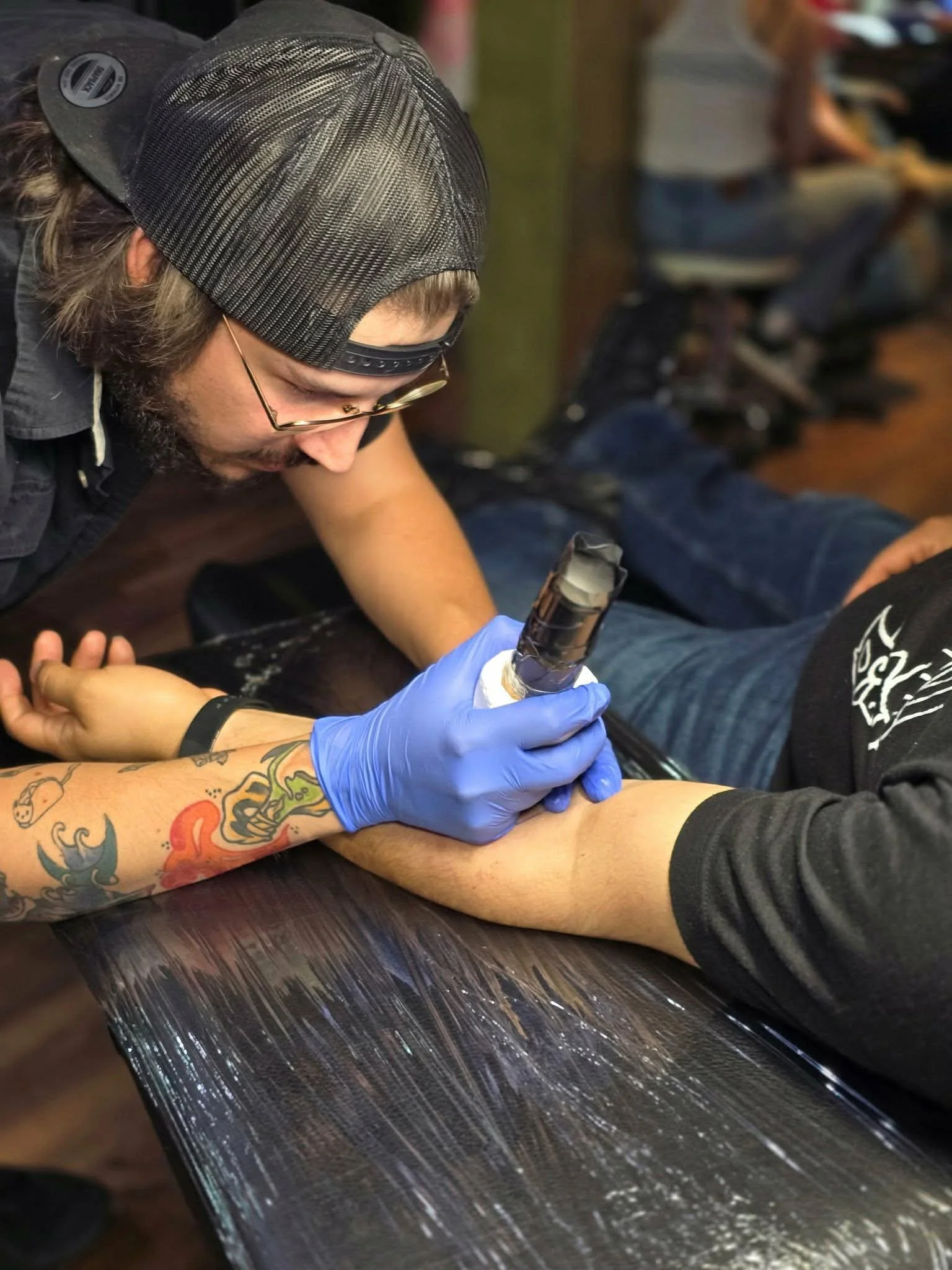 Tattoo artist using a tattoo machine on a person's arm, wearing blue gloves, tattoos visible on the person's arm, in a tattoo studio.