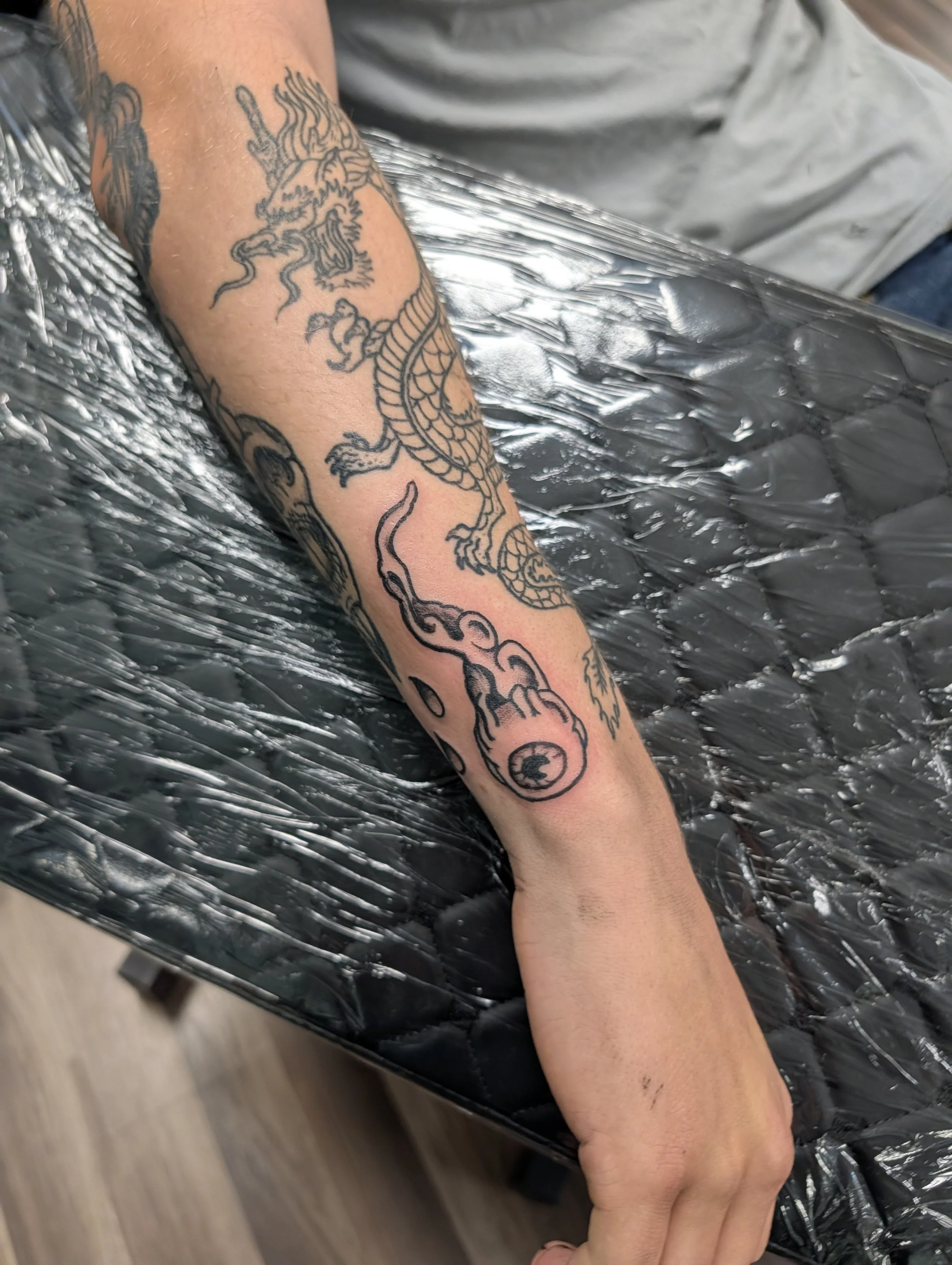 A person's arm with a detailed black tattoo of a dragon, resting on a black plastic-covered surface, likely in a tattoo studio.