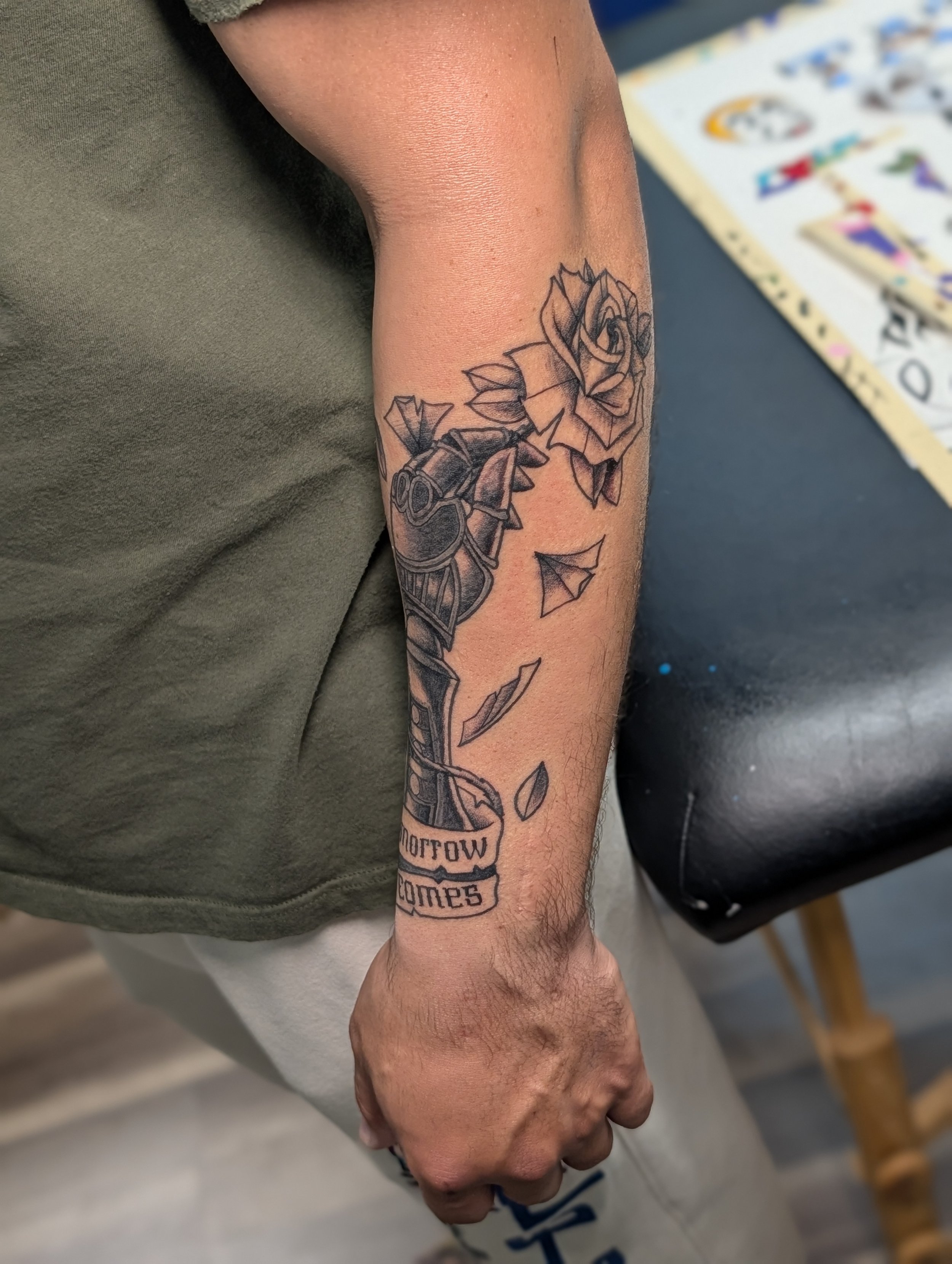 Close-up of a person's forearm with a tattoo of a rose and a handgun, with the words "Tomorrow Comes" written below.