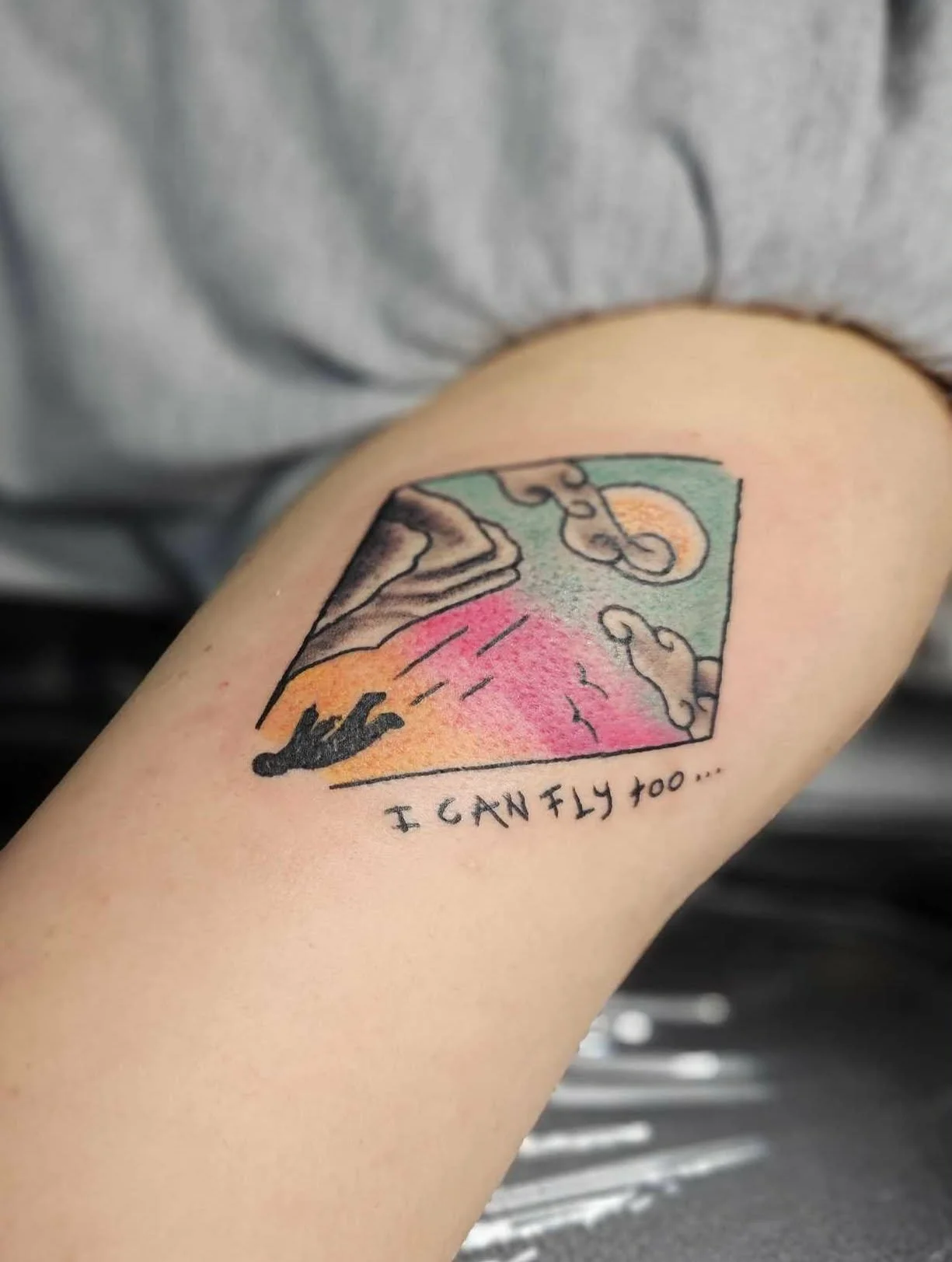 Tattoo of a stylized landscape with a person flying over mountains and clouds, with the text 'I CAN FLY TOO...' beneath it.