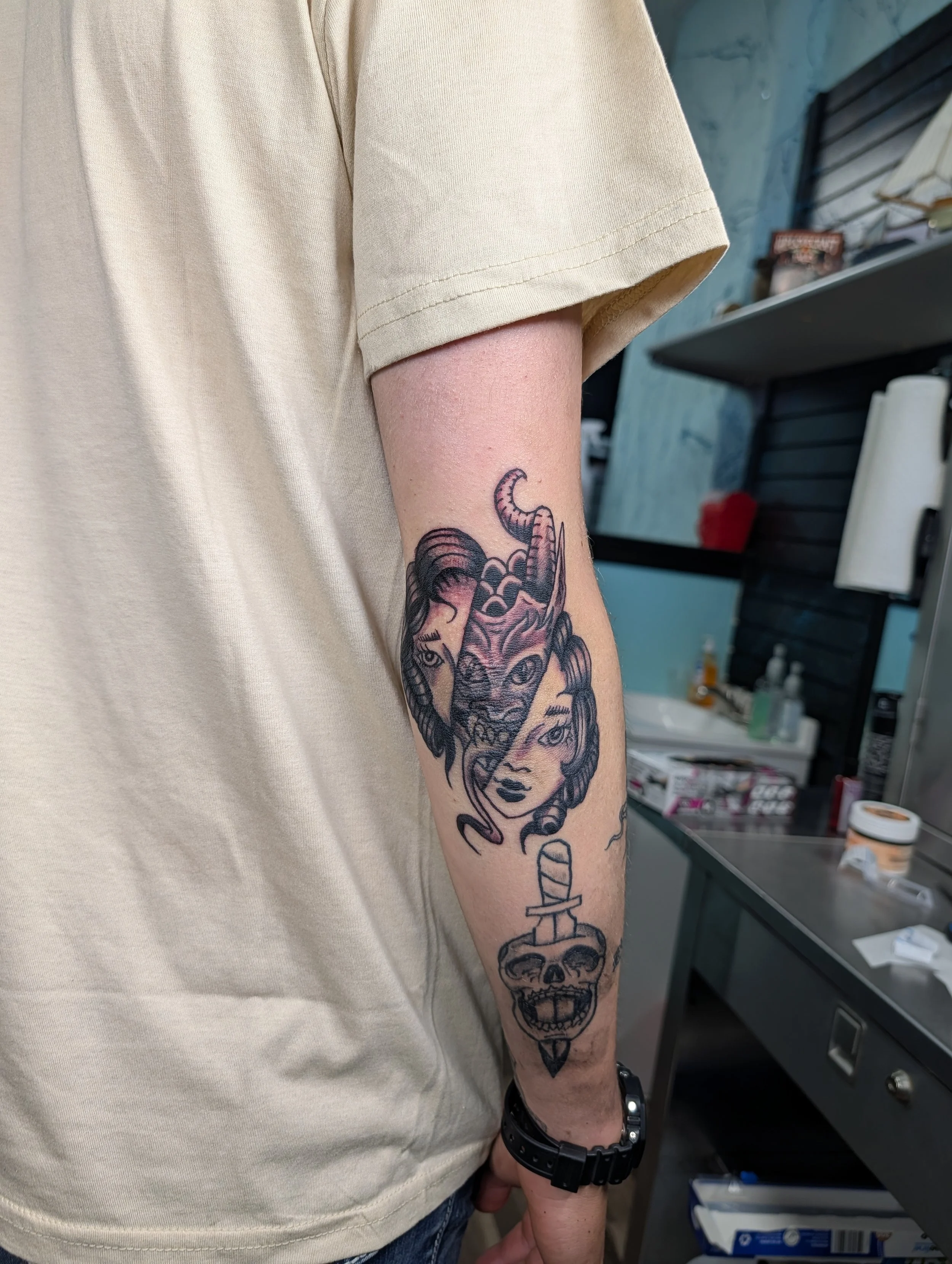A person's arm with multiple tattoos, including a woman’s face, a snake, and a skull, is shown in a tattoo studio or shop.