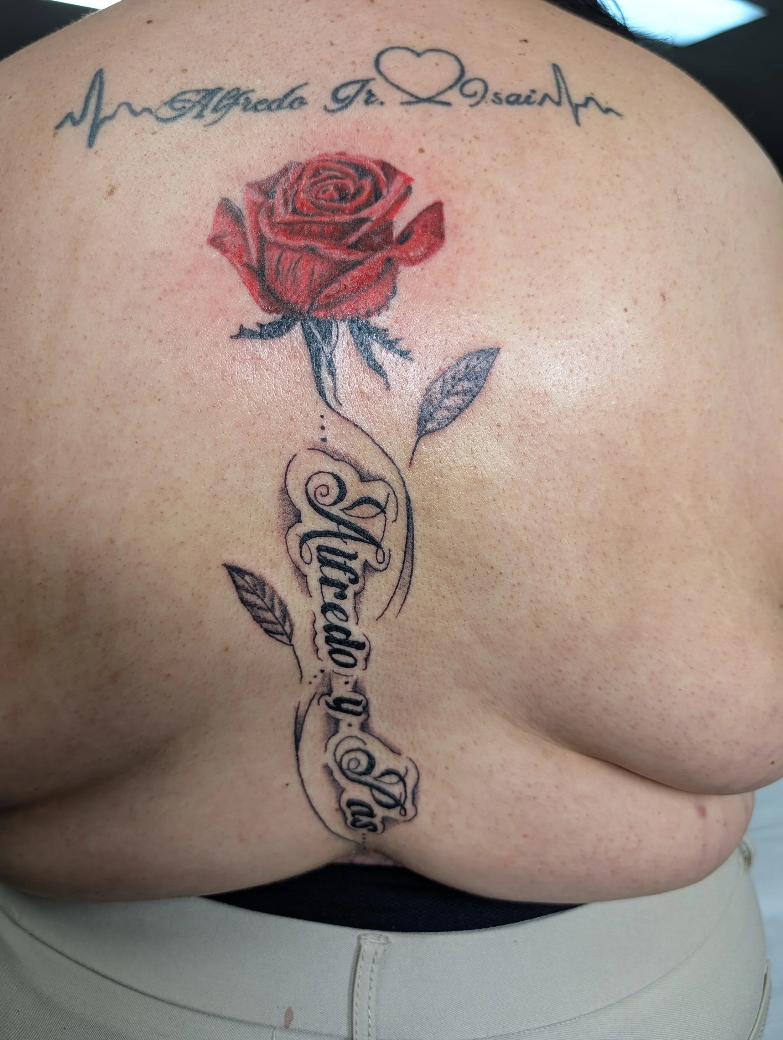 A close-up of a tattoo on a person's upper chest, featuring a red rose, black leaves and stem, and a script phrase with a heart shape.