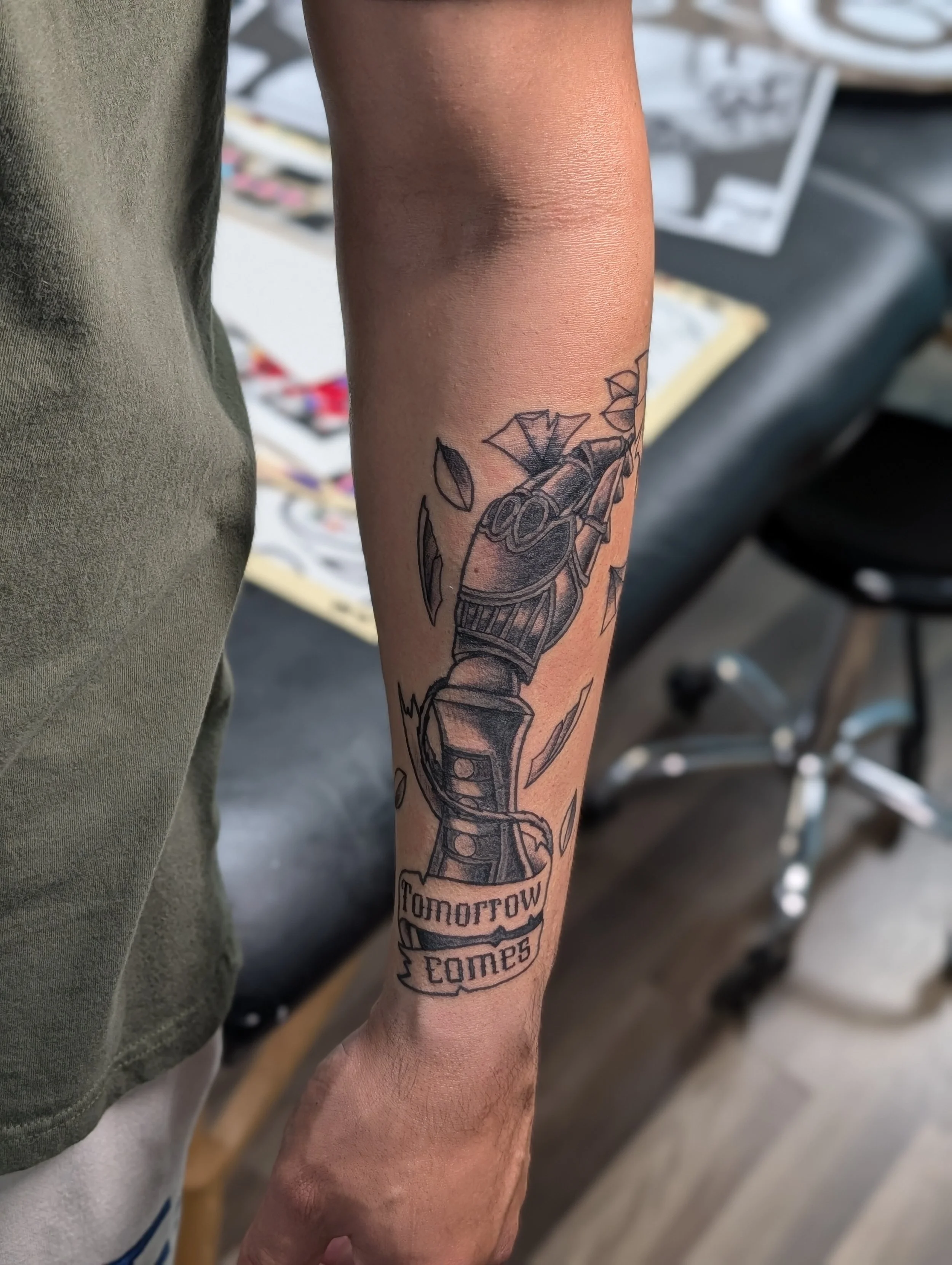 A person's arm with a tattoo of a robot arm holding a sword. The tattoo includes a banner with the words 'tomorrow comes'.