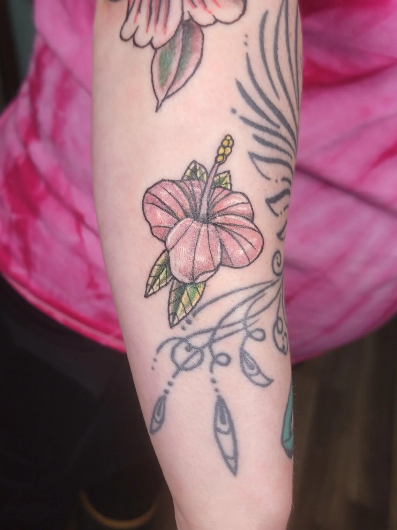 Colorful tattoo of a pink hibiscus flower with green leaves and a yellow stamen on a person's arm, decorated with black outline and tribal-inspired design elements.
