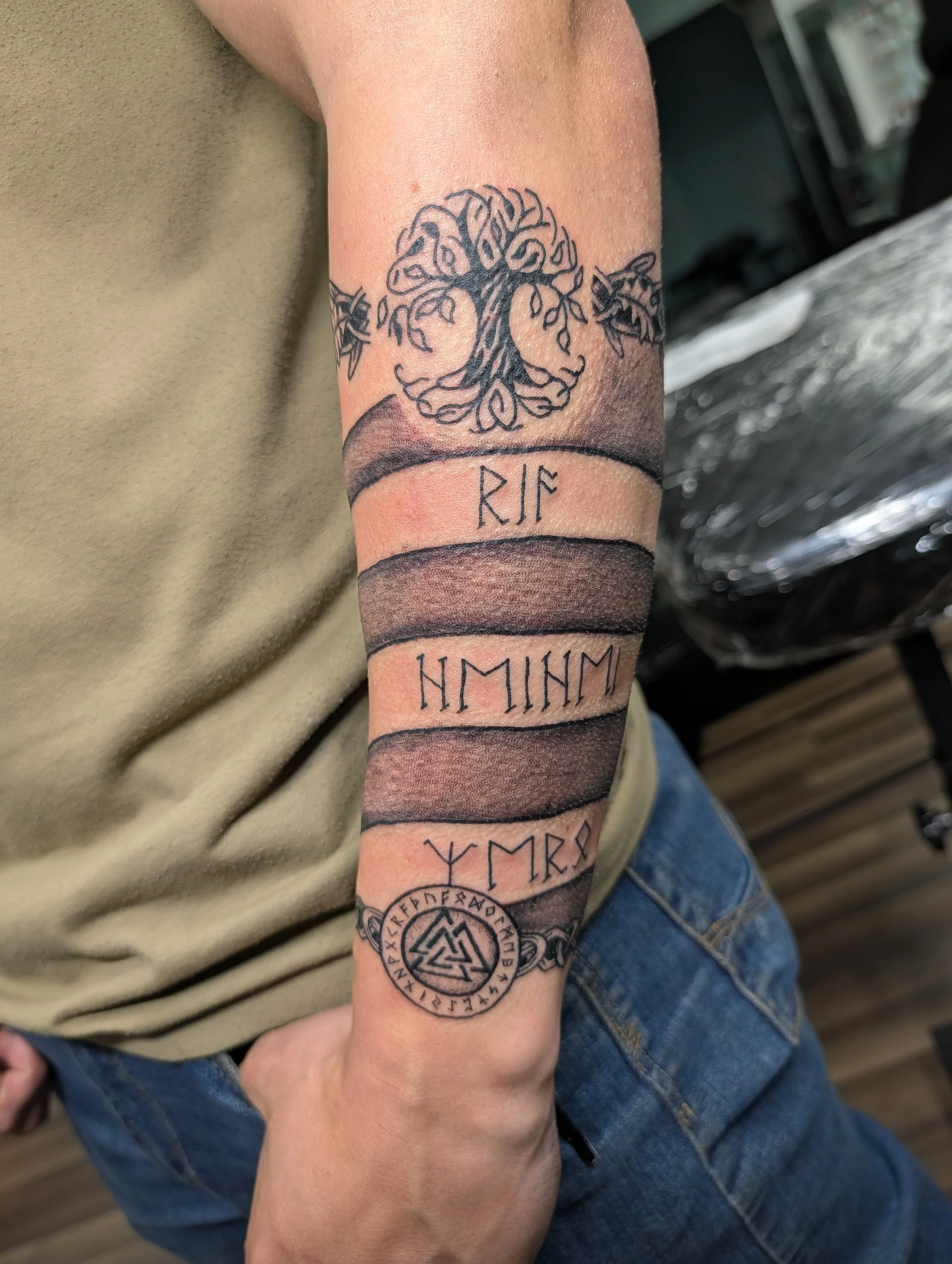 Tattoo of a tree with intertwined roots and branches with an Underworld symbol below, surrounded by runic inscriptions on a person's forearm.
