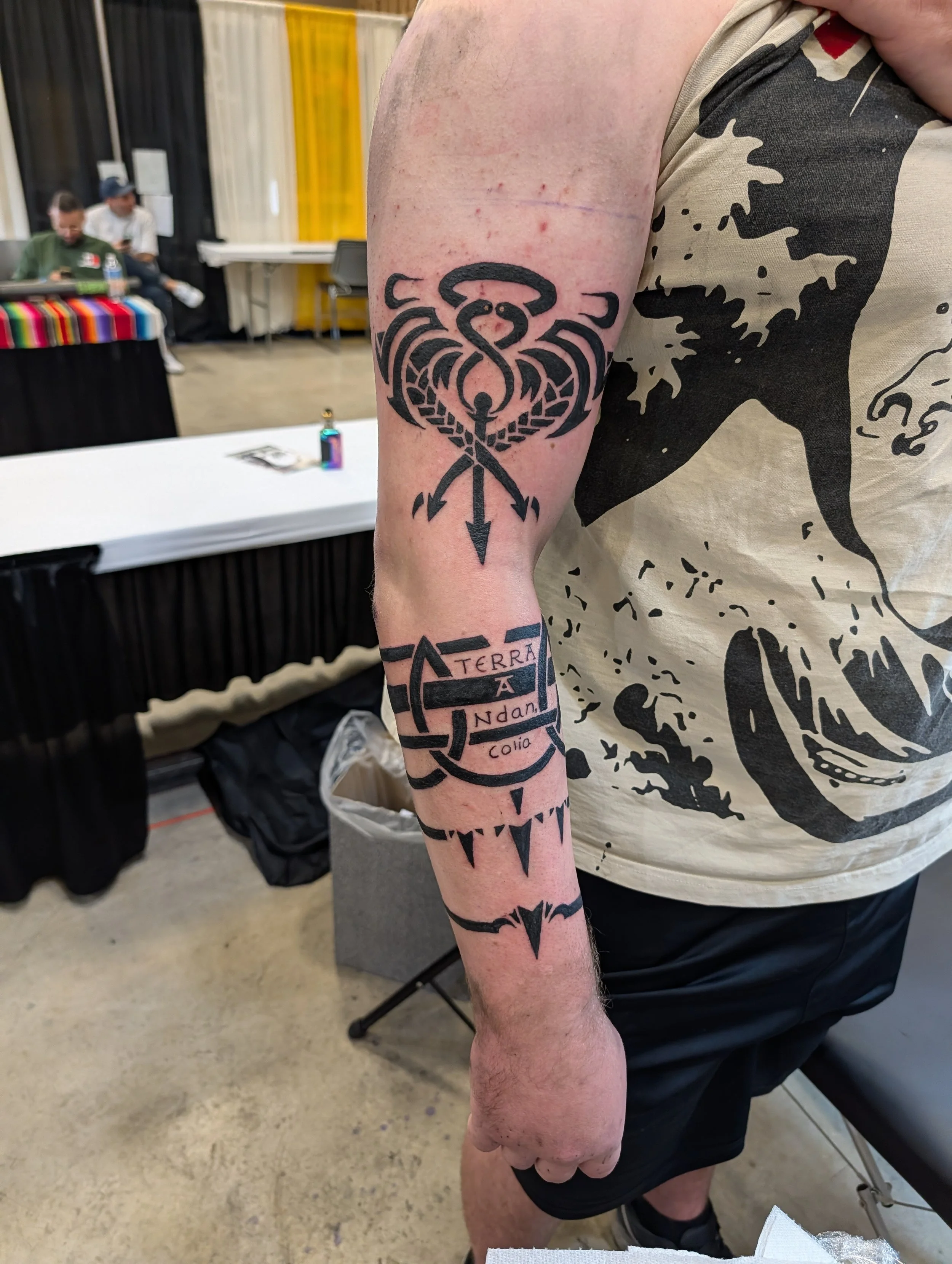 Tattoo on person's upper arm featuring a nautical star with arrows pointing downward, and some words including 'TERRA,' 'A,' 'NDAN,' and 'Colo' in smaller font.