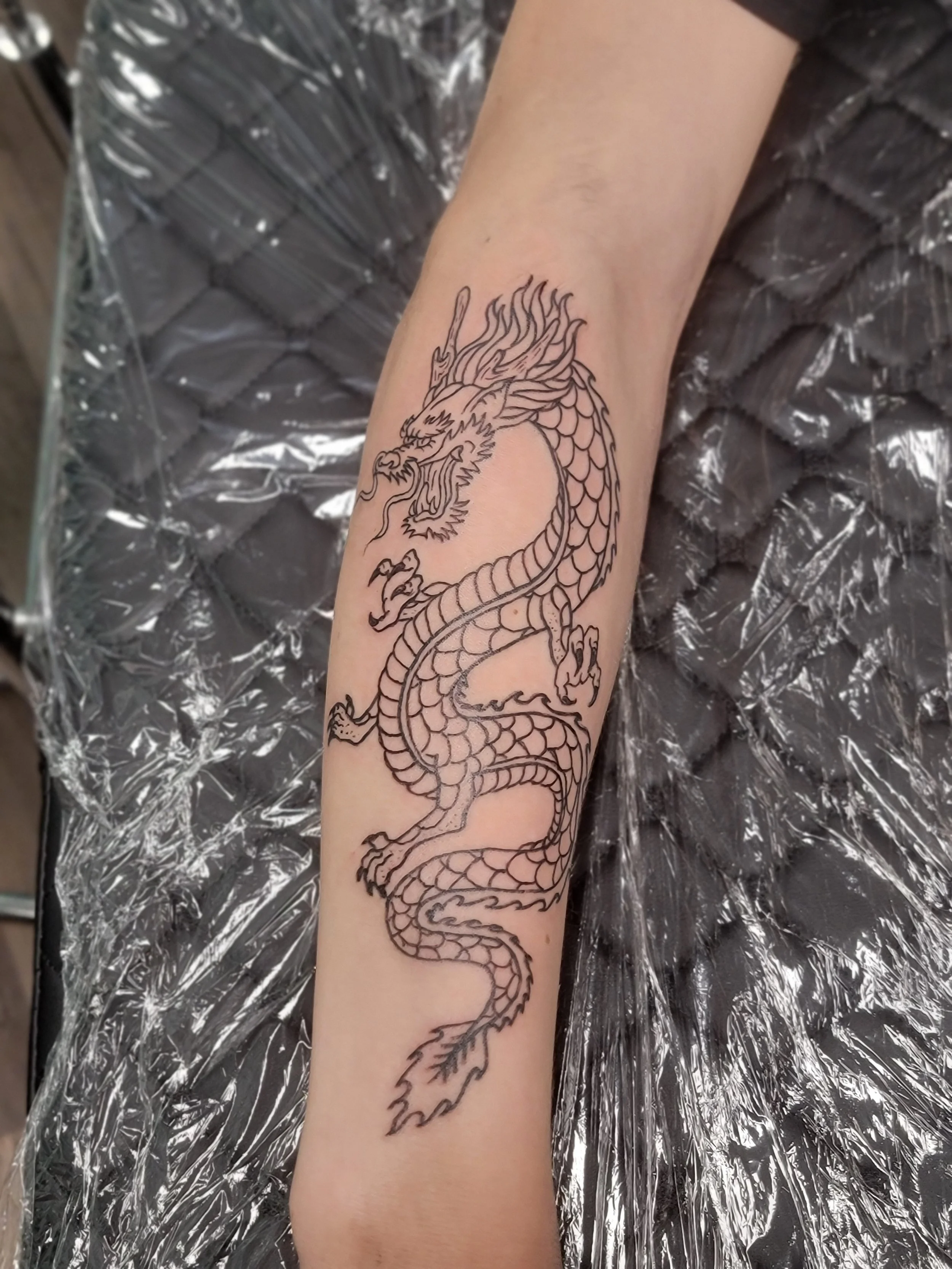 A detailed black outline of a traditional Chinese dragon tattoo on a person's forearm.