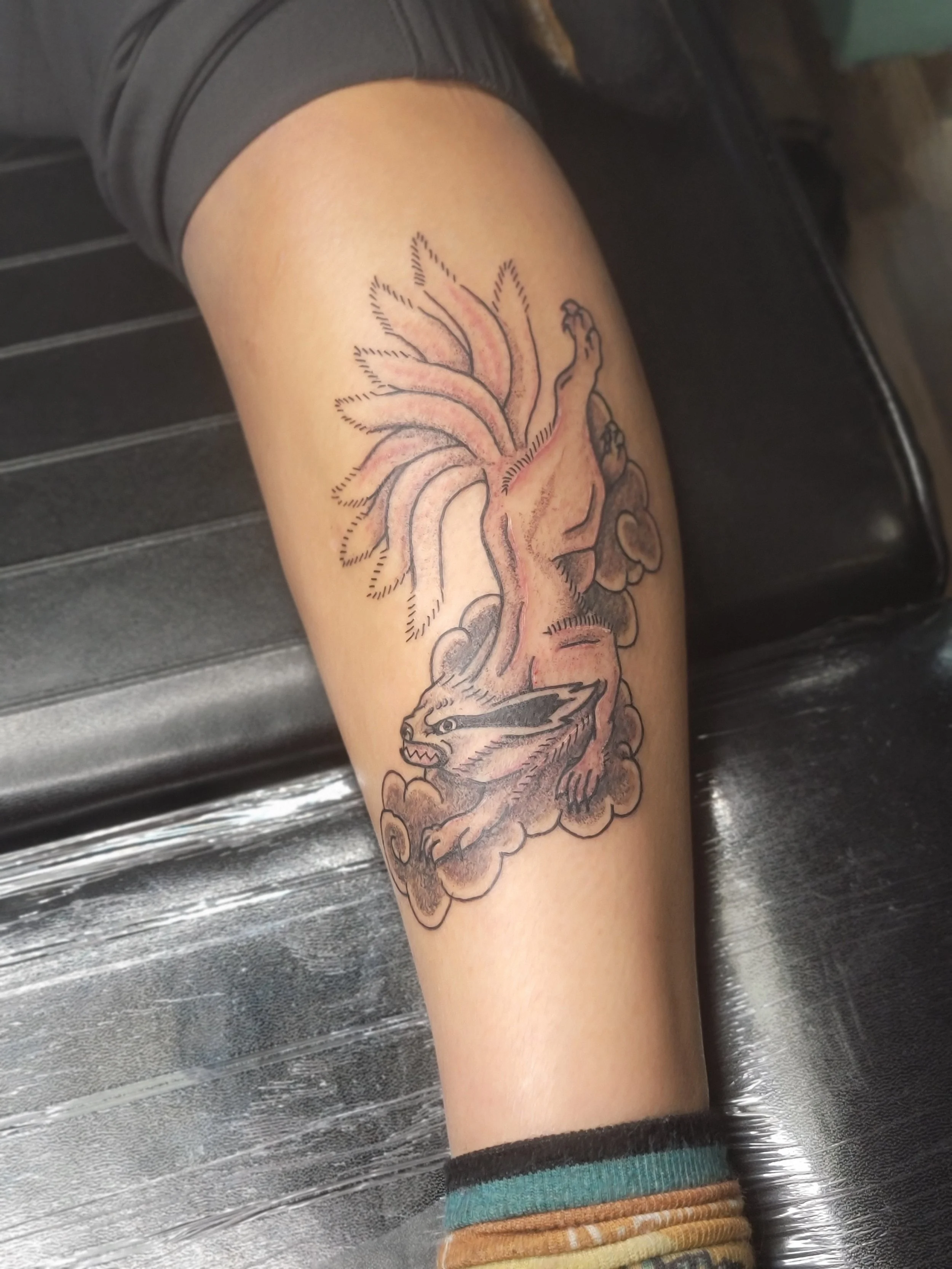 Tattoo of a wolf intertwined with a tree on a person's arm, with clouds at the bottom and colorful bracelets.