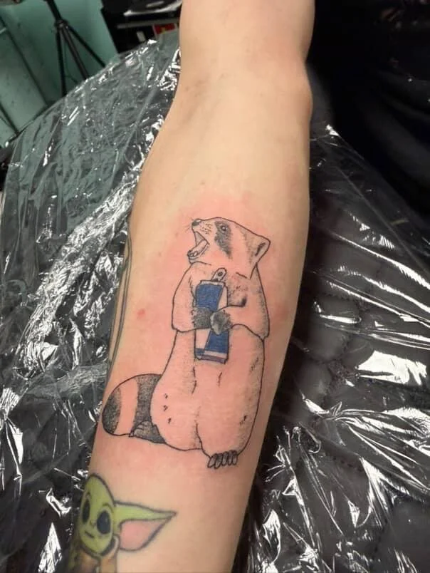 Tattoo of raccoon holding a redbull with its mouth open, on a person's forearm.