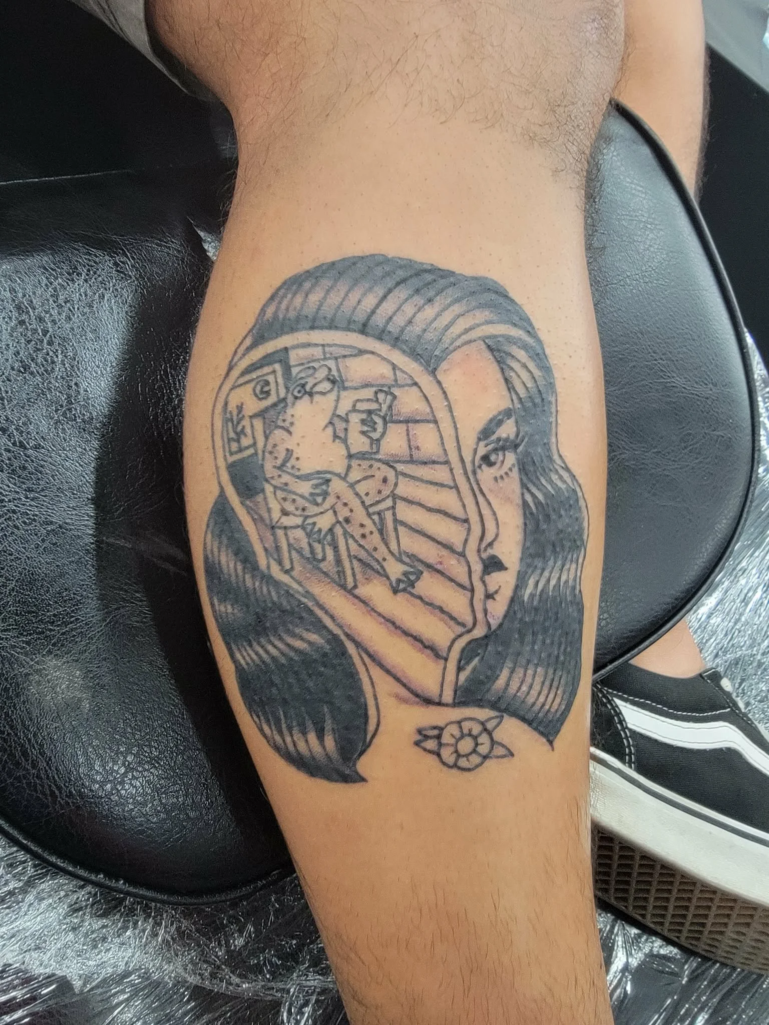 Tattoo of a woman's face with long hair, containing an image of a frog sitting on a chair inside her hairline, holding a drink, with a flower below.