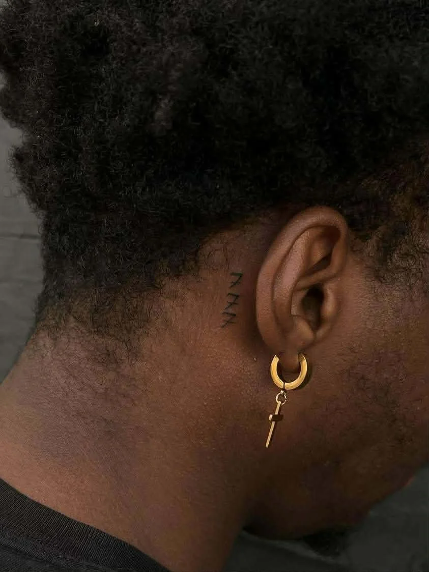 Close-up of a person's ear with a gold hoop earring that has a cross pendant. Behind the ear, there is a tattoo of three small black '7's.