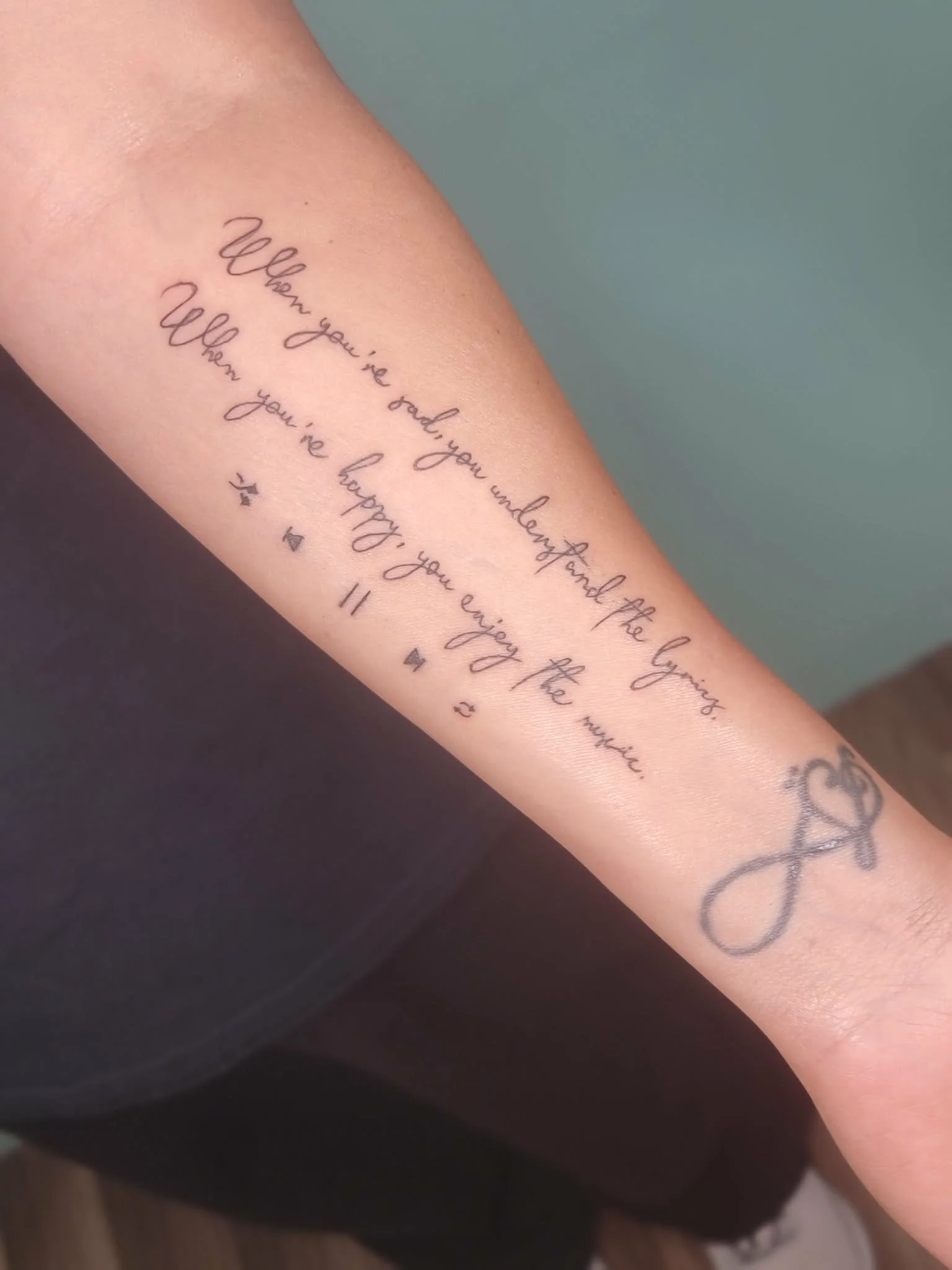 A tattoo on a person's forearm with a quote in cursive script that reads, 'When you're sad, you understand the feeling, when you're happy, you enjoy the moment,' with small decorative stars and an infinity symbol at the end.