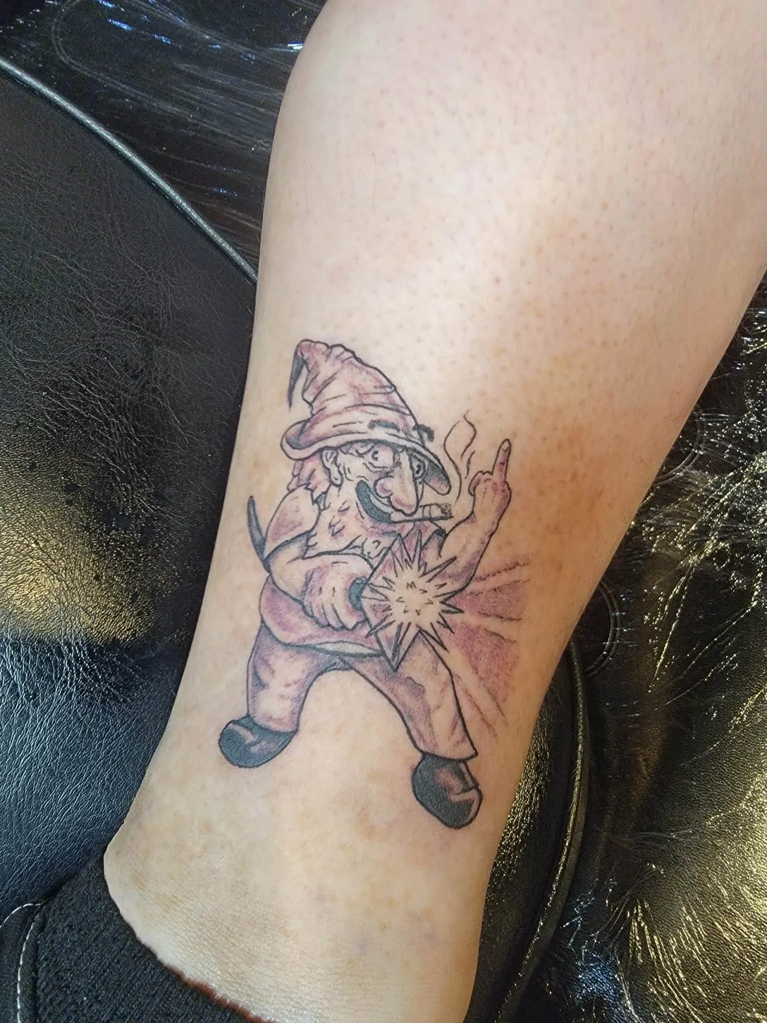 Tattoo of a gnome wearing a hat, holding a lit bomb, and giving an obscene gesture with his middle finger.
