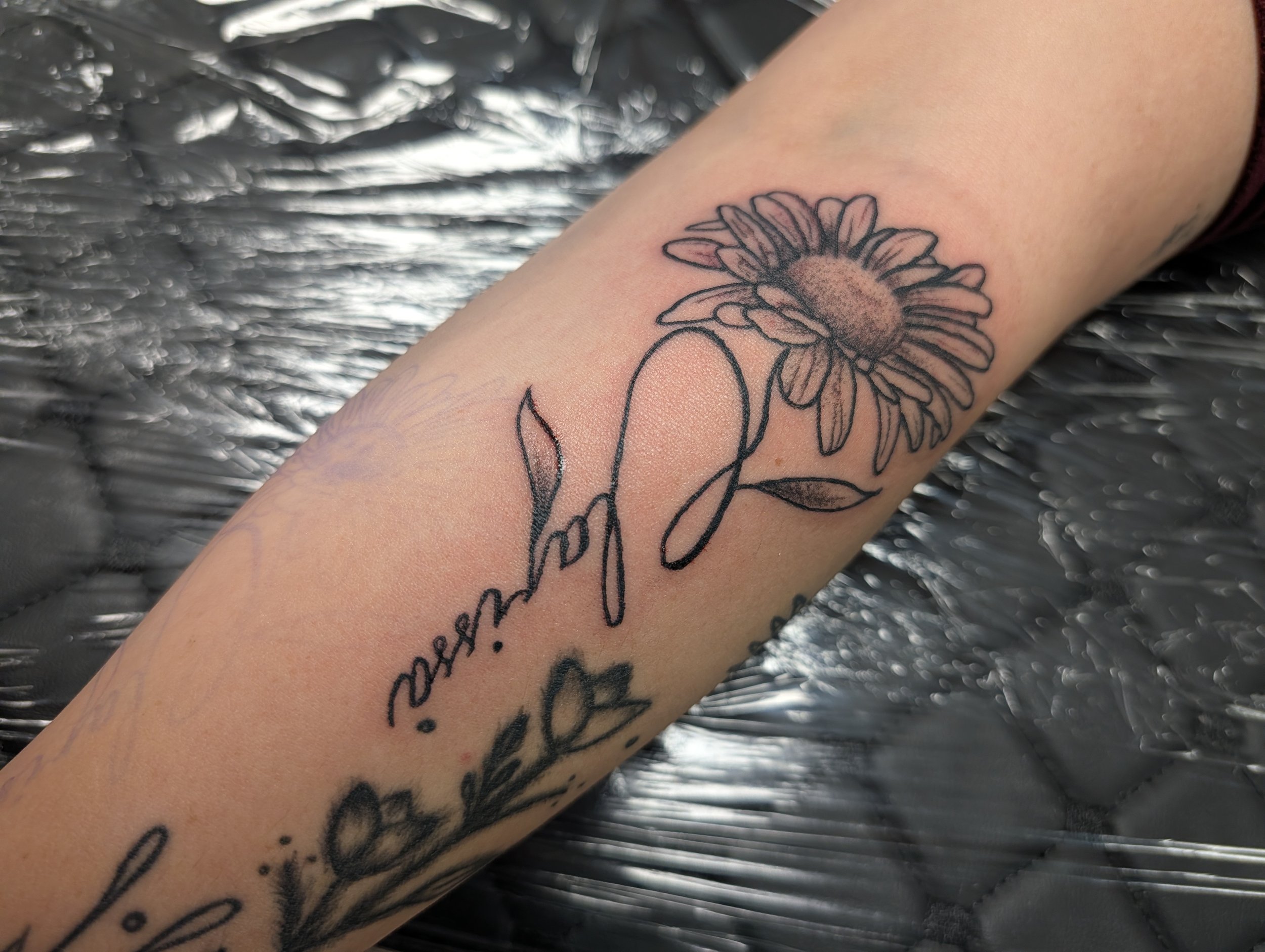 A tattoo of a daisy flower with a shaded center on a person's forearm, with cursive writing below the flower. The background is a crinkled, metallic surface.