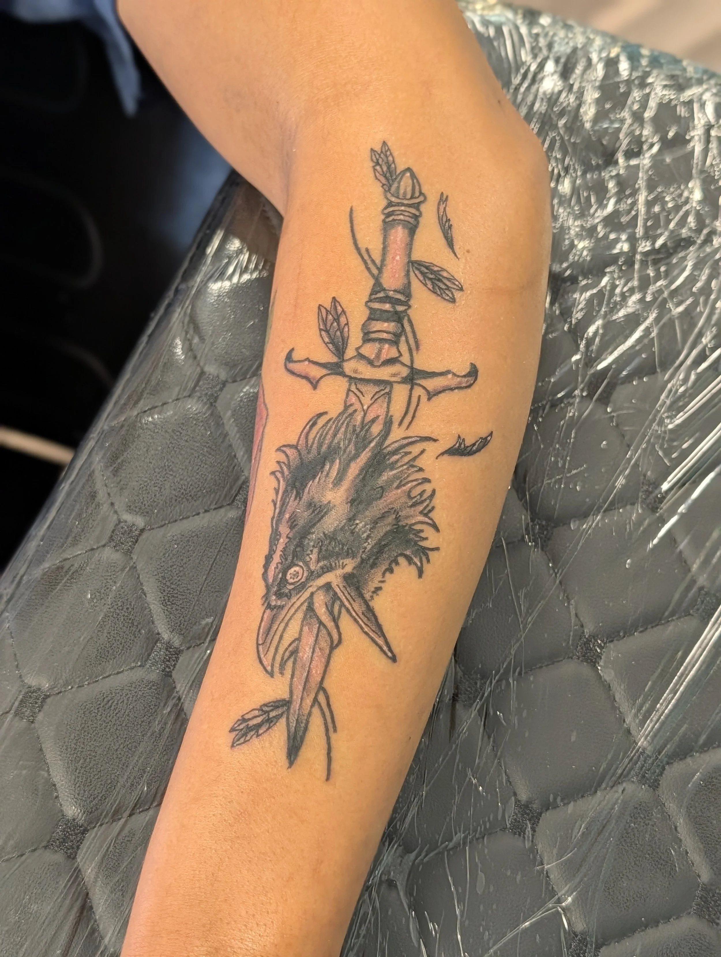 A tattoo of a sword with leaves on the arm, with a lion's face visible below it, on a person's forearm.