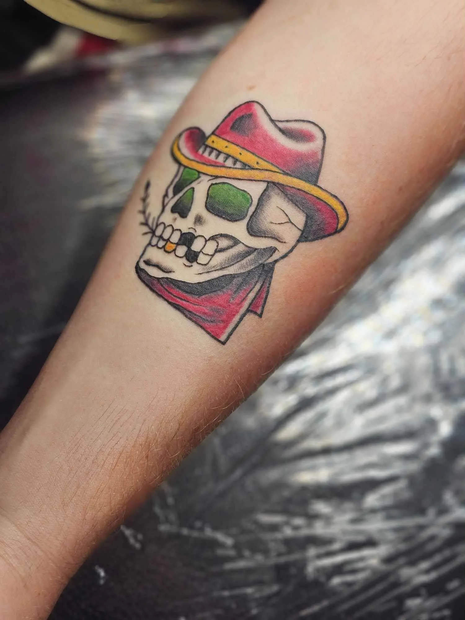 Tattoo of a skull wearing a red cowboy hat, green eye sockets, and a red bandana.