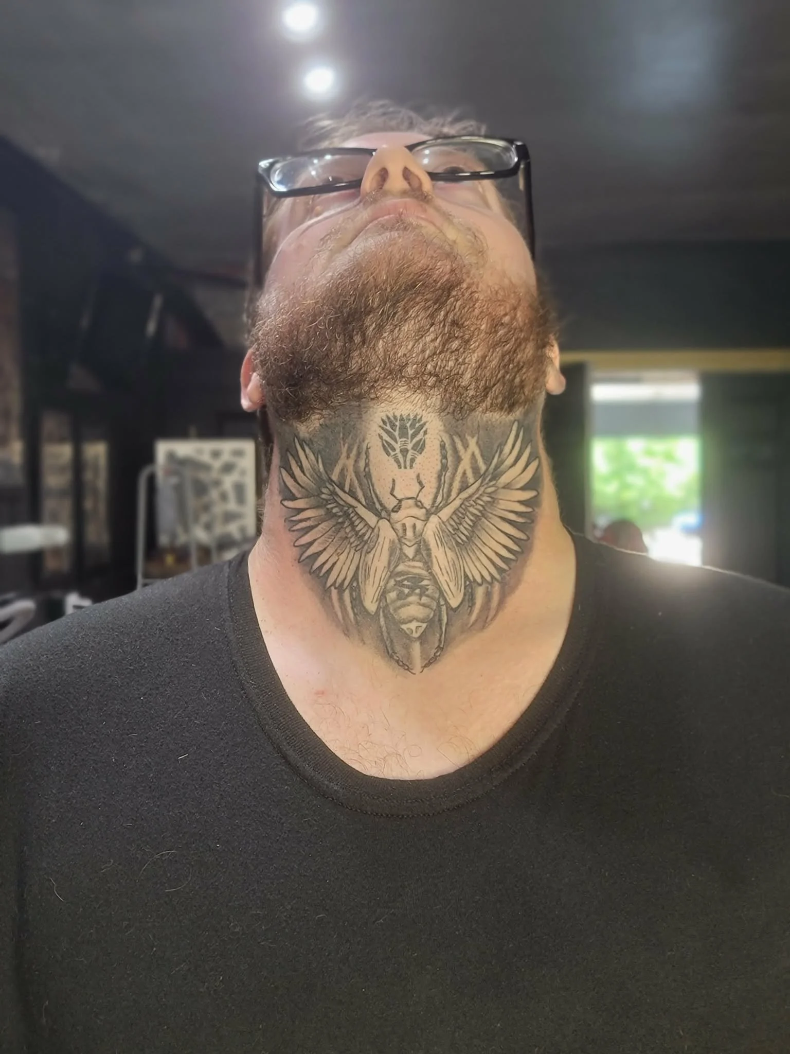 A man with a beard and glasses, looking upward. He has a large tattoo on his neck featuring a scarab beetle with outstretched wings and a geometric design above it.