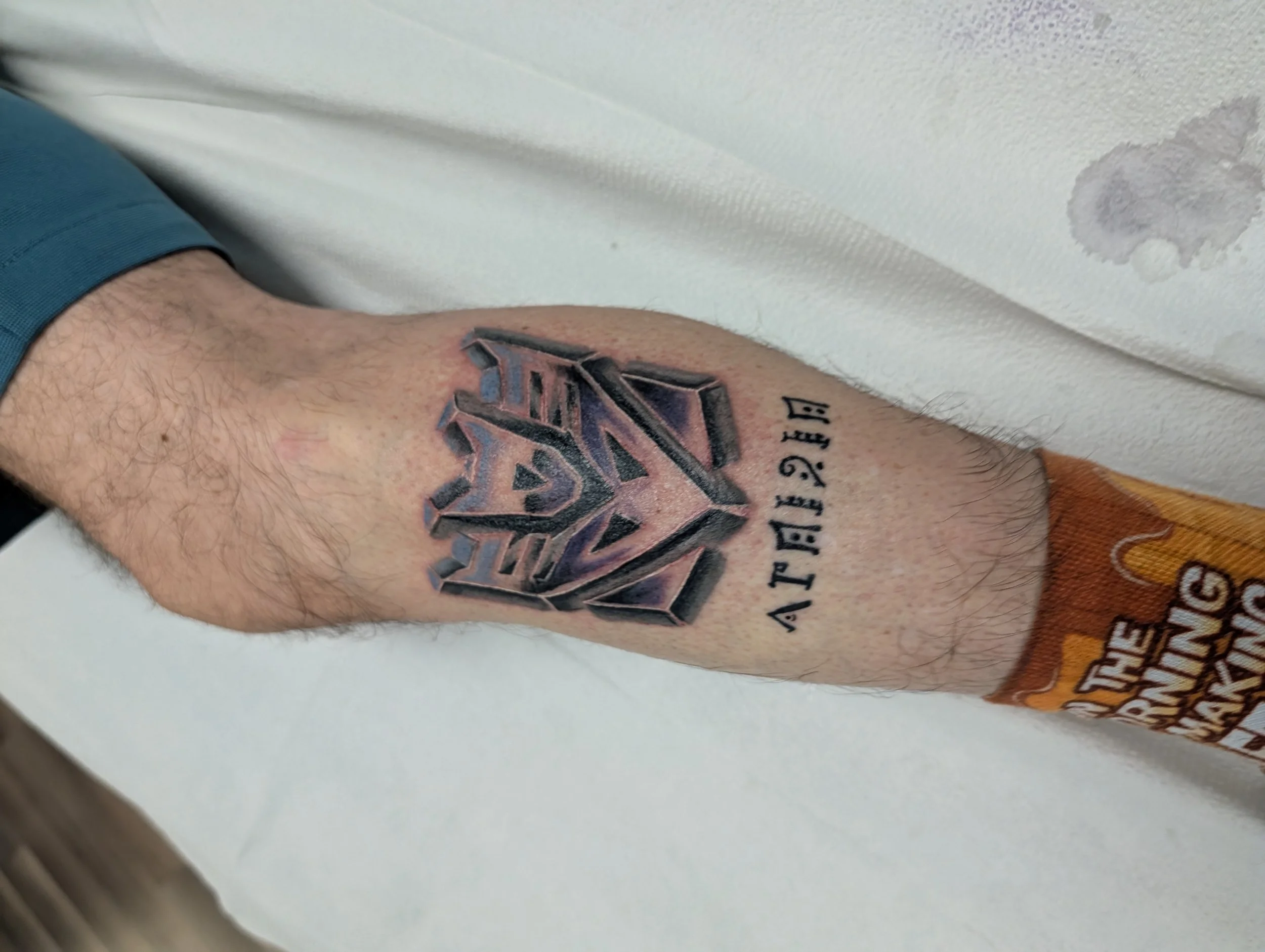 Close-up photo of a person's arm with a colorful tattoo of a geometric design, and part of a shirt sleeve visible. The background includes a white textured surface with a water stain.