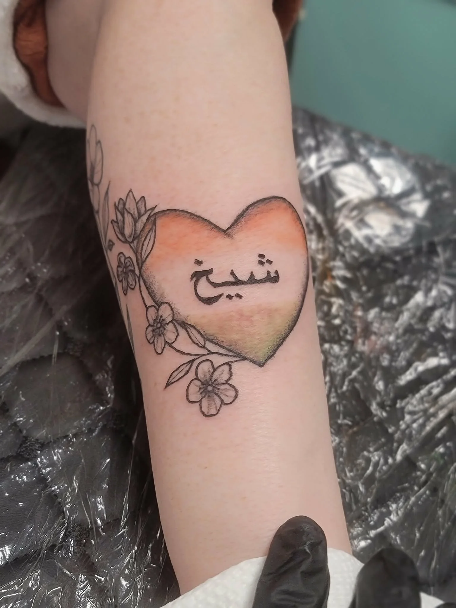 A tattoo of a heart with a colored gradient, surrounded by flowers and leaves, with Arabic script inside the heart.