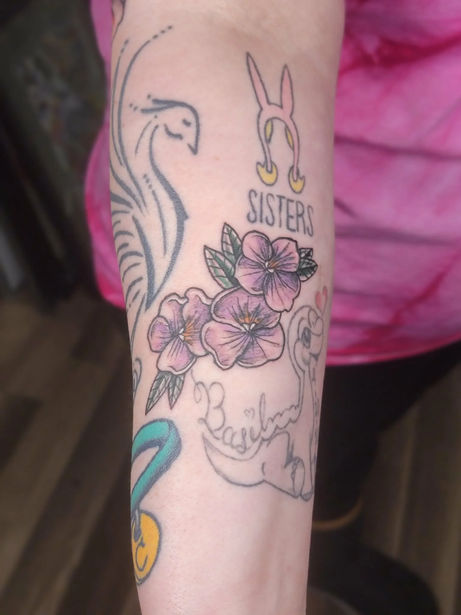 Tattoo on forearm featuring floral design, a banner with the word "sisters," and other decorative elements.