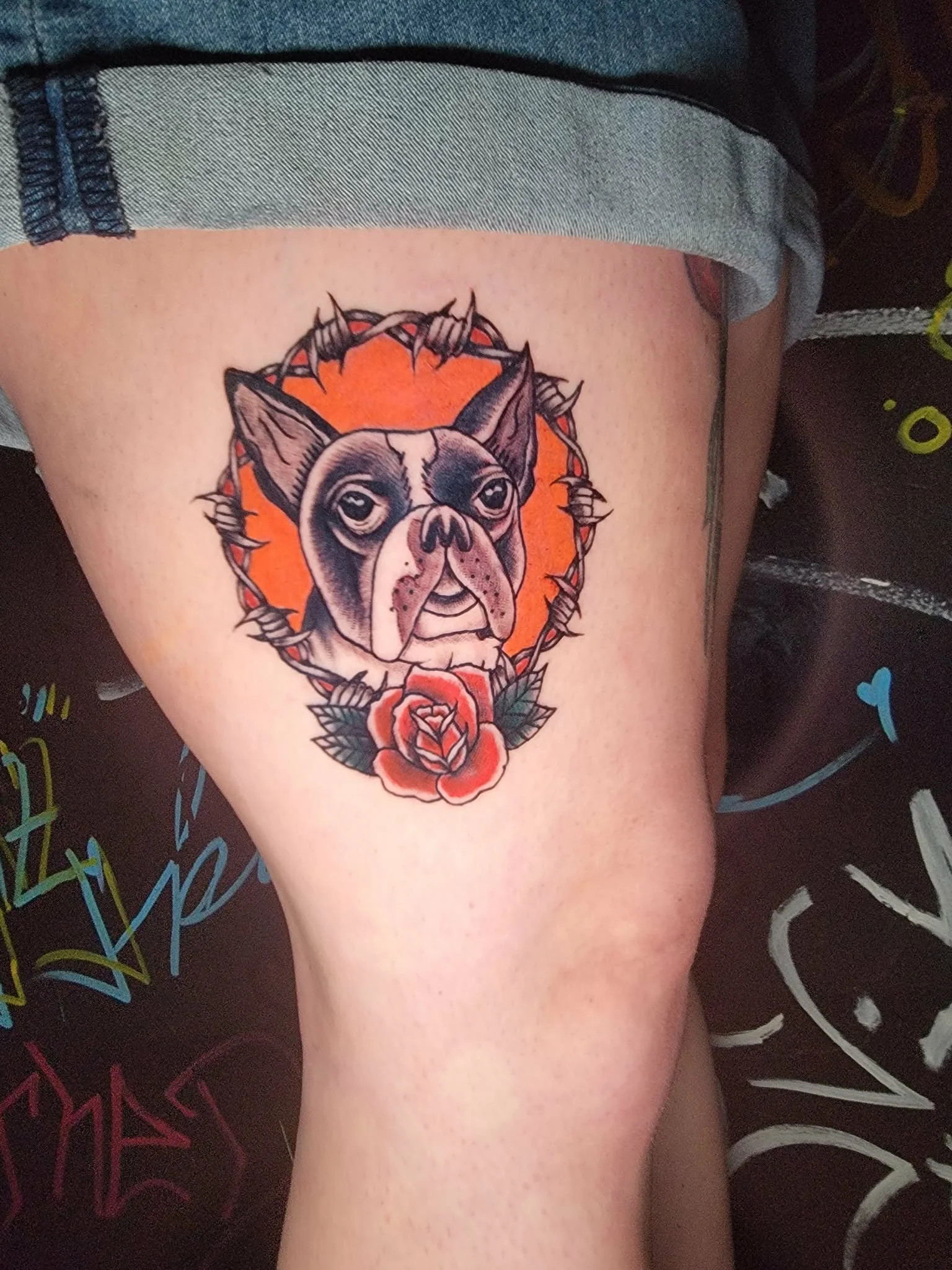 Tattoo of a dog's head, possibly a Boston Terrier, within a thorny circular frame. Below it, a red rose with green leaves. The background behind the dog is orange.