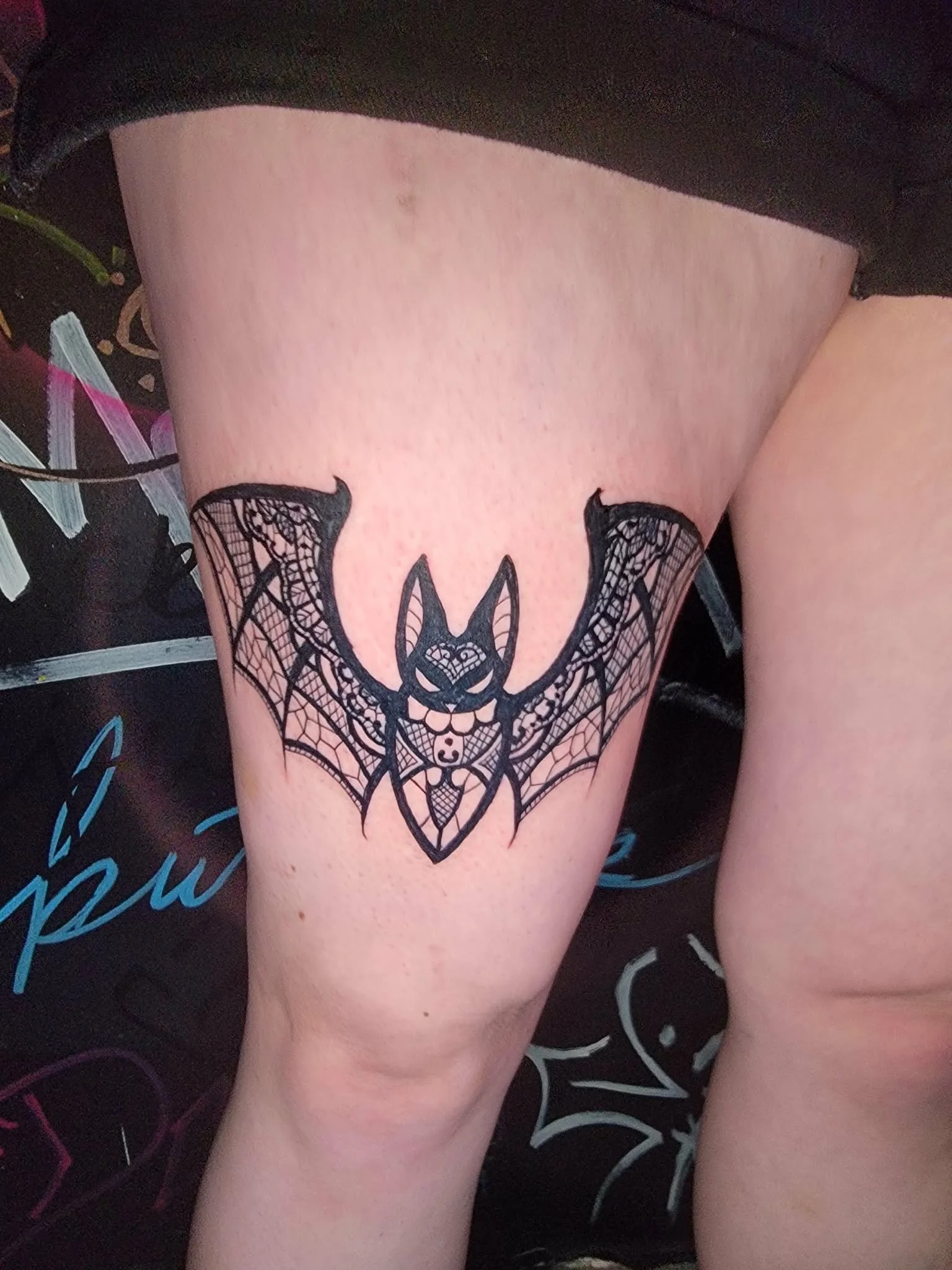 Black tattoo of a bat with a cat-like face on a person's thigh.