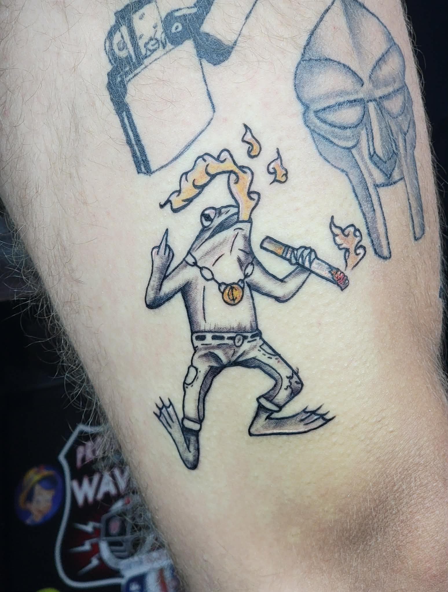 A tattoo artwork depicting a cartoon frog character smoking a cigar, wearing baggy pants, a necklace with a pendant, and sunglasses, with a confident pose.