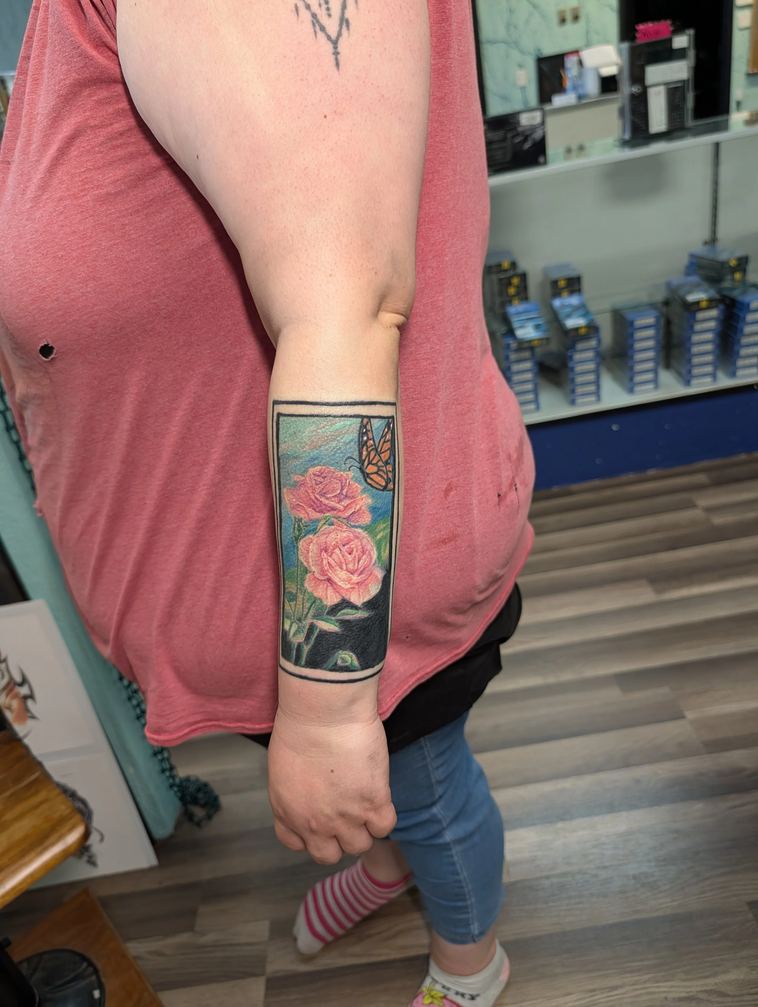 A person wearing a pinkish-red T-shirt, blue jeans, striped socks, and white shoes with a butterfly design. The person has tattoos on the forearm and upper arm, including a colorful floral design with a butterfly. In the background, there are shelves