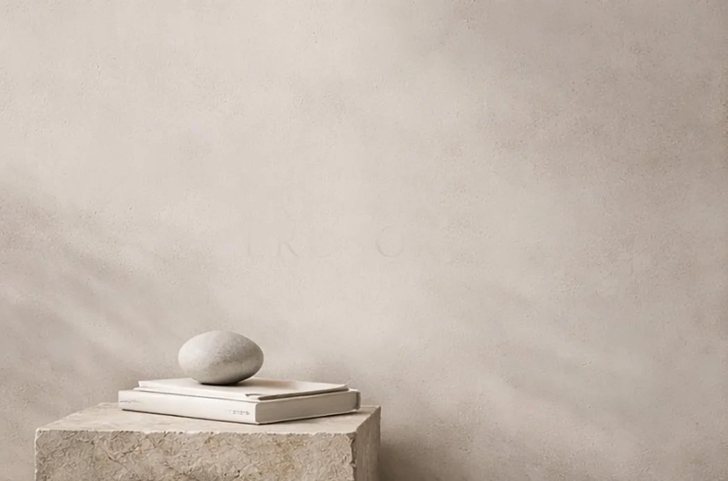 Minimalist arrangement of a rounded stone on top of a few stacked books, placed on a textured stone pedestal against a plain wall.