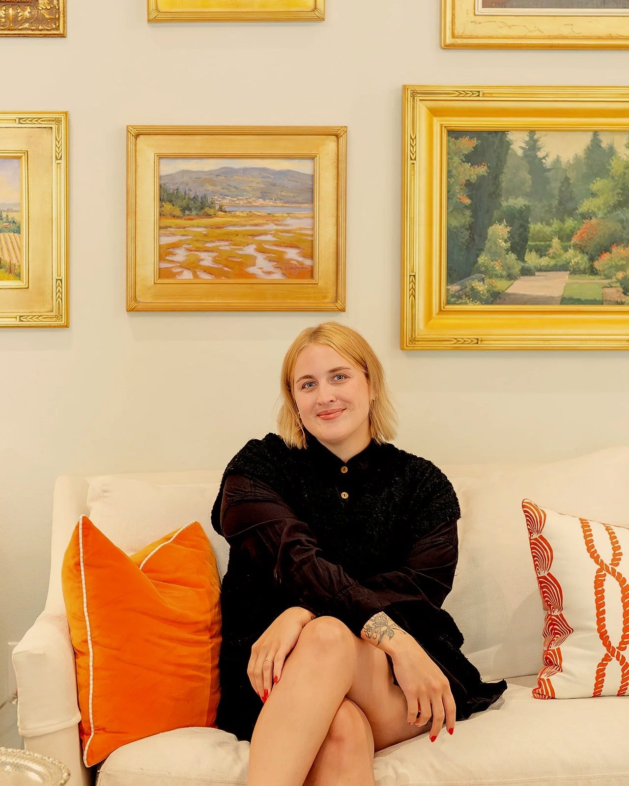 Young woman with blonde hair sitting on a white sofa in an art gallery, surrounded by framed landscape paintings and decorative pillows in orange and white.