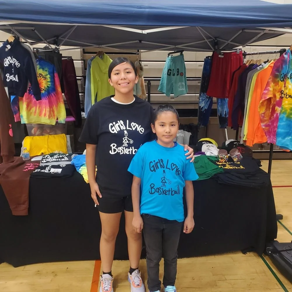 Two young girls standing in front of a booth with colorful basketball-themed apparel, including T-shirts, shorts, and hoodies, at an indoor sports event. The girls are wearing T-shirts that say "Girls Love Basketball."