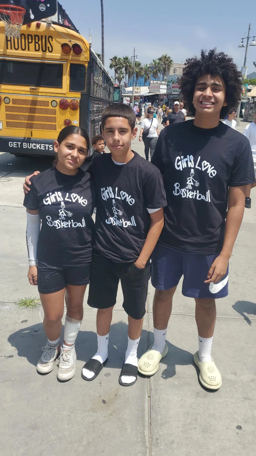 Three young individuals standing outdoors, wearing matching black t-shirts with white text that says, "Girls Love Basketball," with a small stick figure holding a basketball graphic. They are smiling and posing for the photo. In the background, there