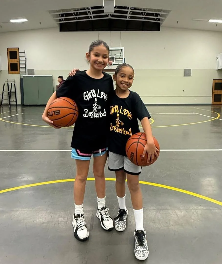 Two young girls smiling and posing for a photo on a basketball court. They are wearing black t-shirts with 'Girls Love Basketball' printed on them, and each is holding a basketball. The court is indoors with a basketball hoop in the background.
